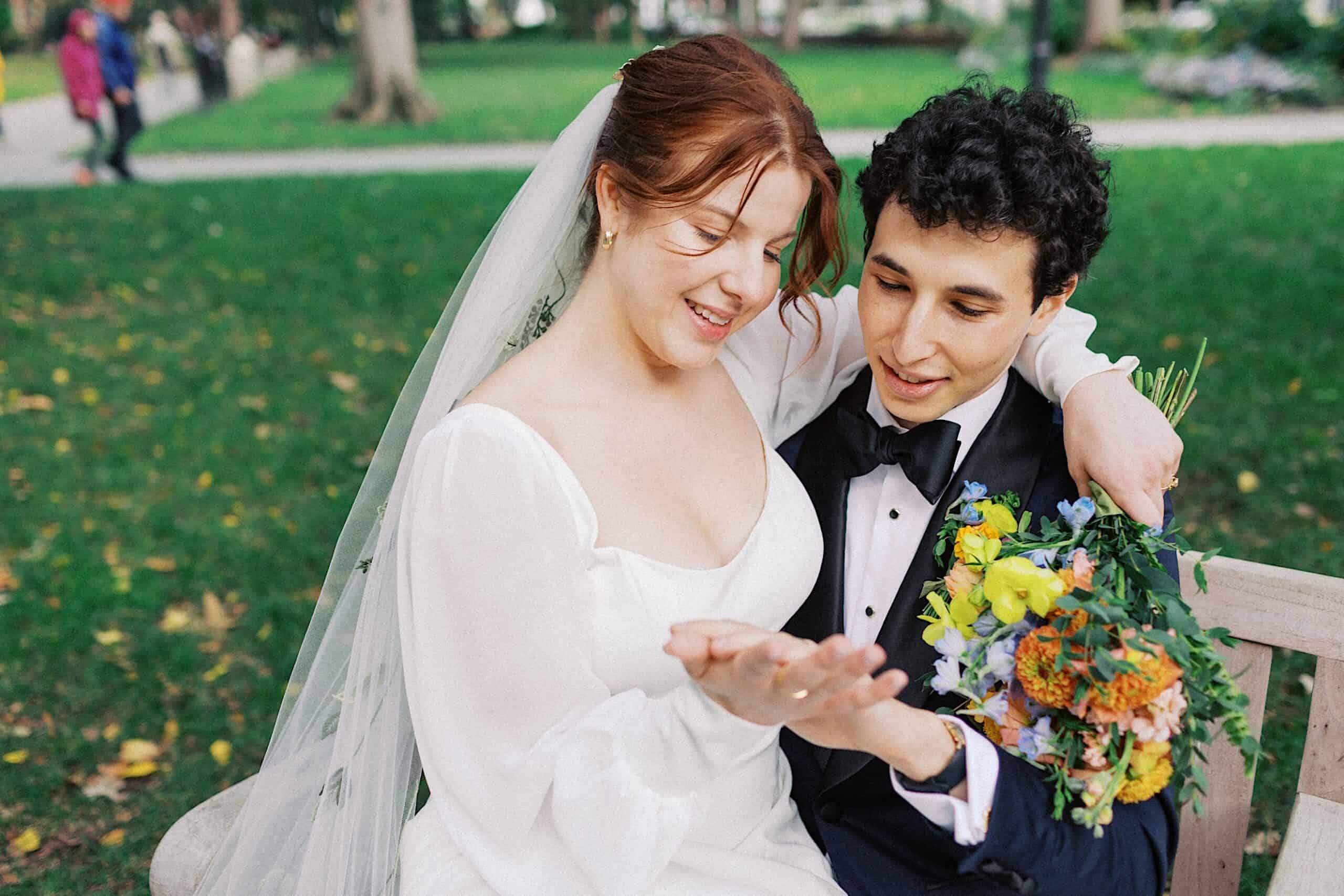 A bride and groom sit on a bench in a park. The bride smiles and shows her hand, while the groom holds a bouquet of flowers and looks at her hand.