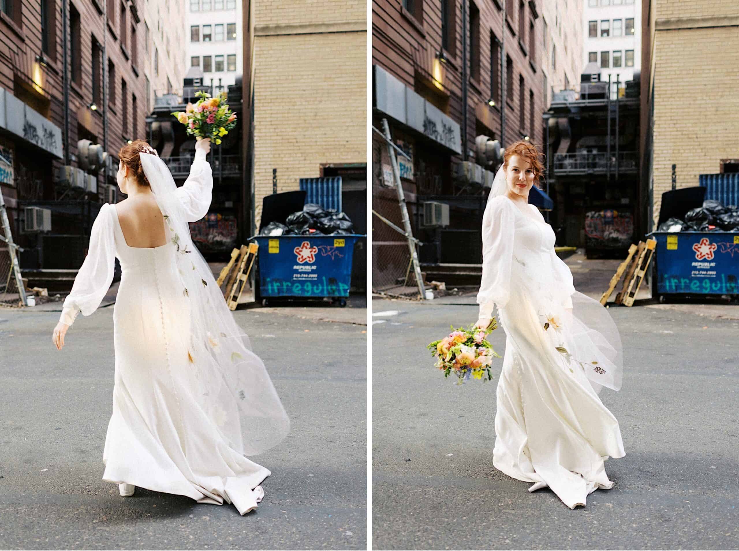 A bride in a long white dress and veil holds a bouquet while standing in an urban alley with graffiti, dumpsters, and tall buildings in the background.