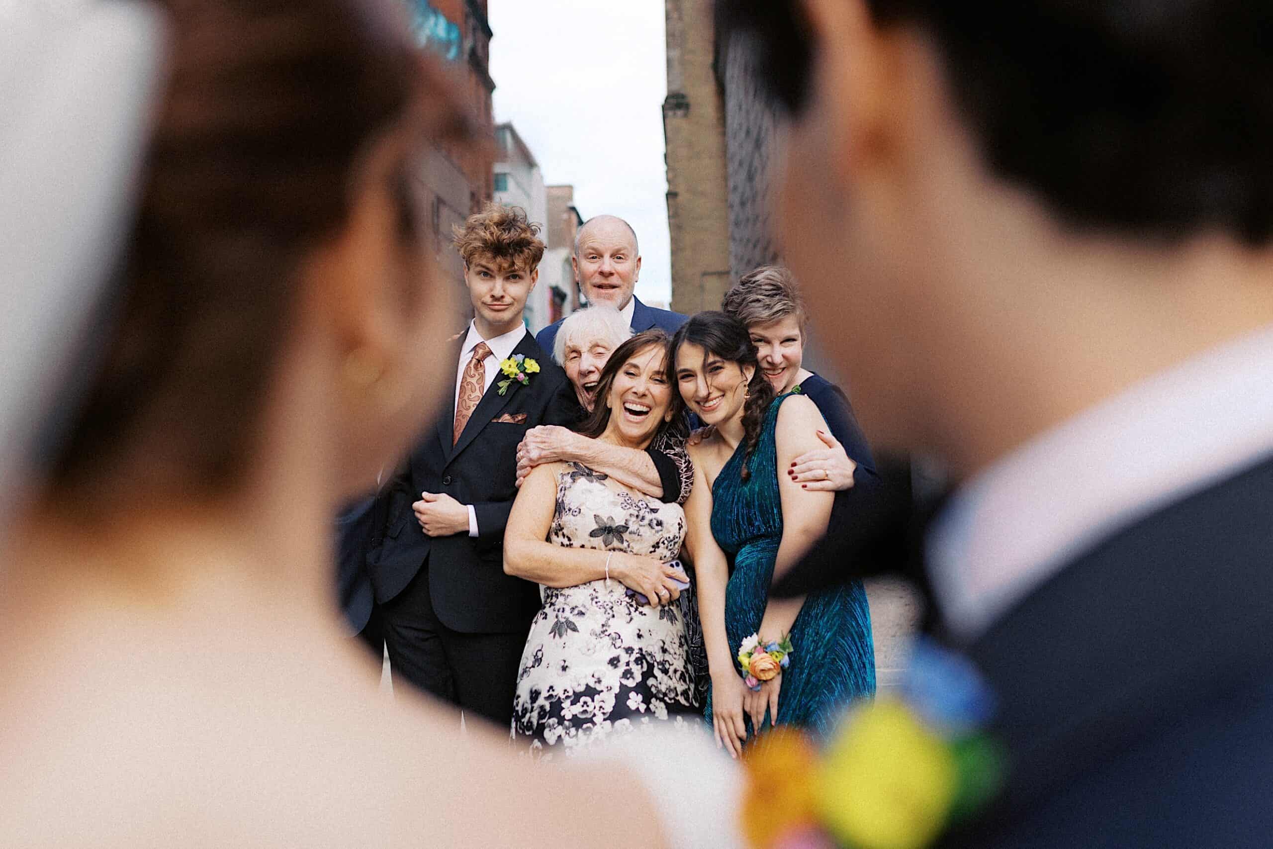 A group of people, dressed formally, smile and pose for a photo outdoors, framed by the blurred silhouettes of two people in the foreground.