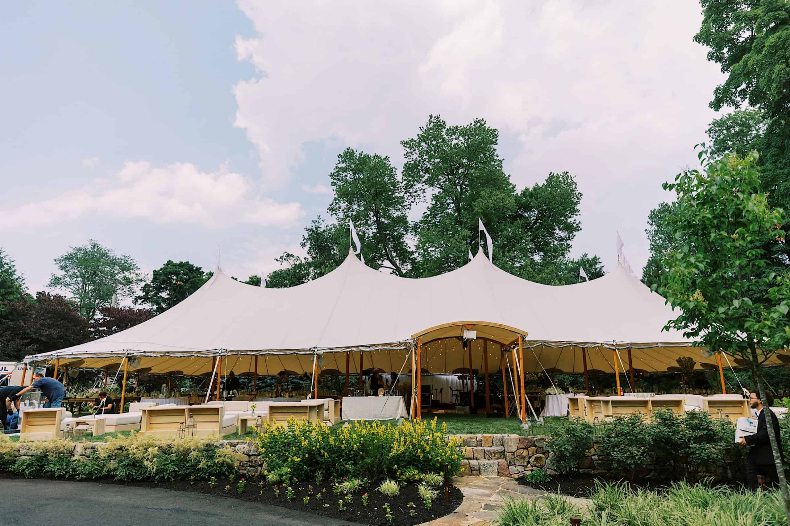 Large white event tent with open sides set up outdoors for a romantic backyard wedding in NJ, surrounded by greenery, flowers, and wooden seating arrangements.