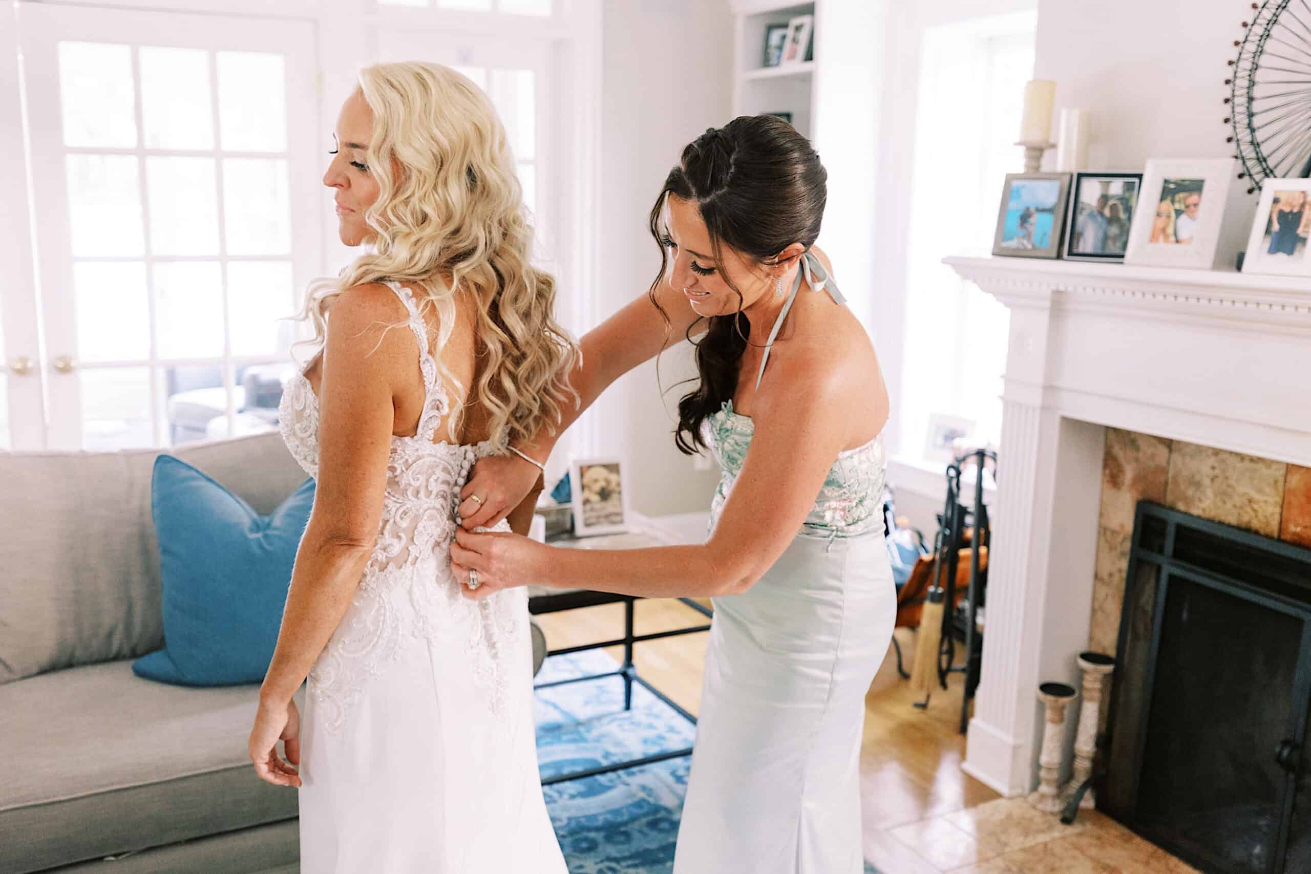 Two women in formal dresses stand in a living room; one adjusts the back of the other’s white dress, possibly preparing for a romantic backyard wedding in NJ.