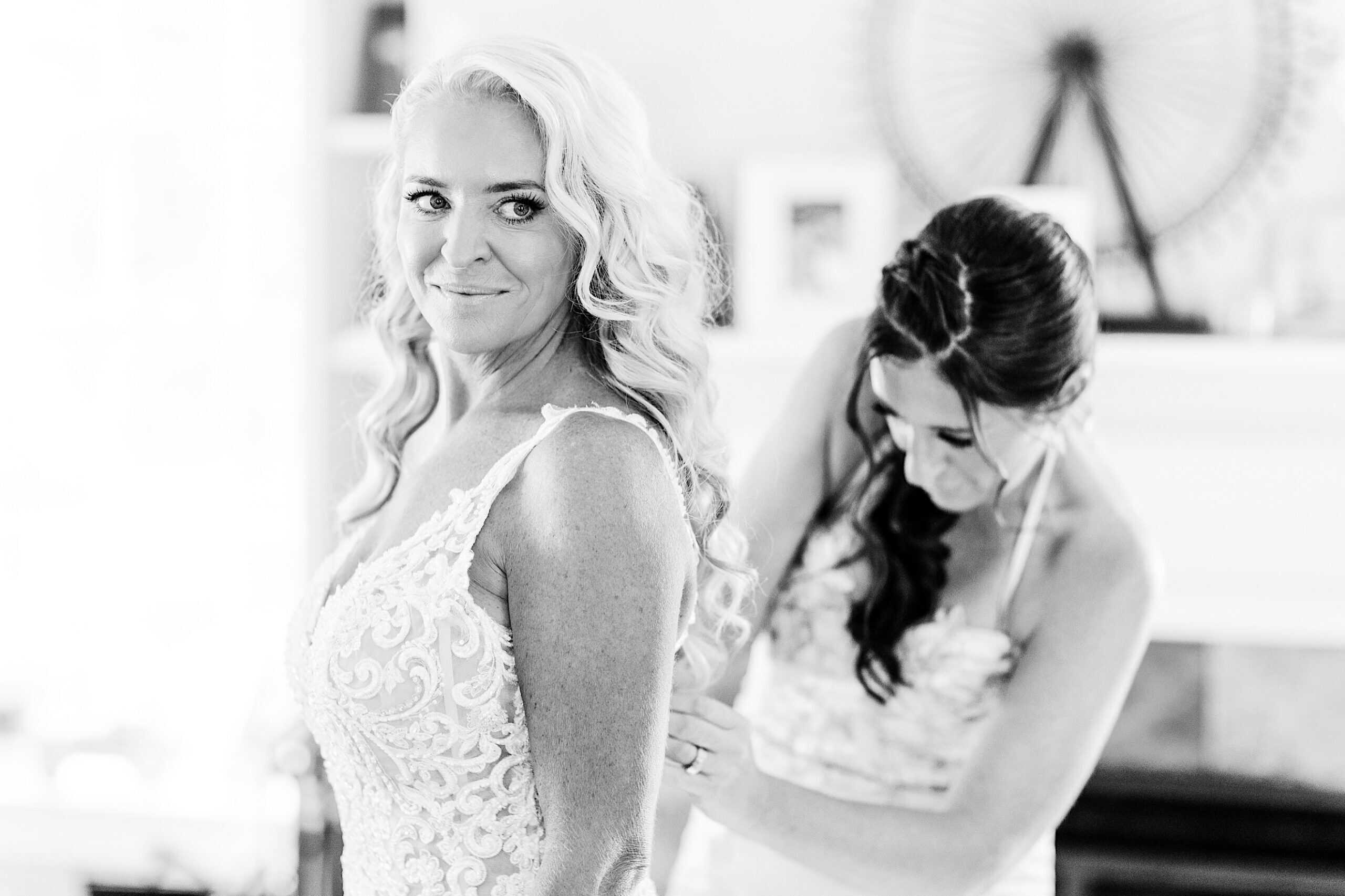 A woman in a wedding dress stands smiling while another woman helps fasten the back of her dress in a bright room, preparing for a romantic backyard wedding in NJ.