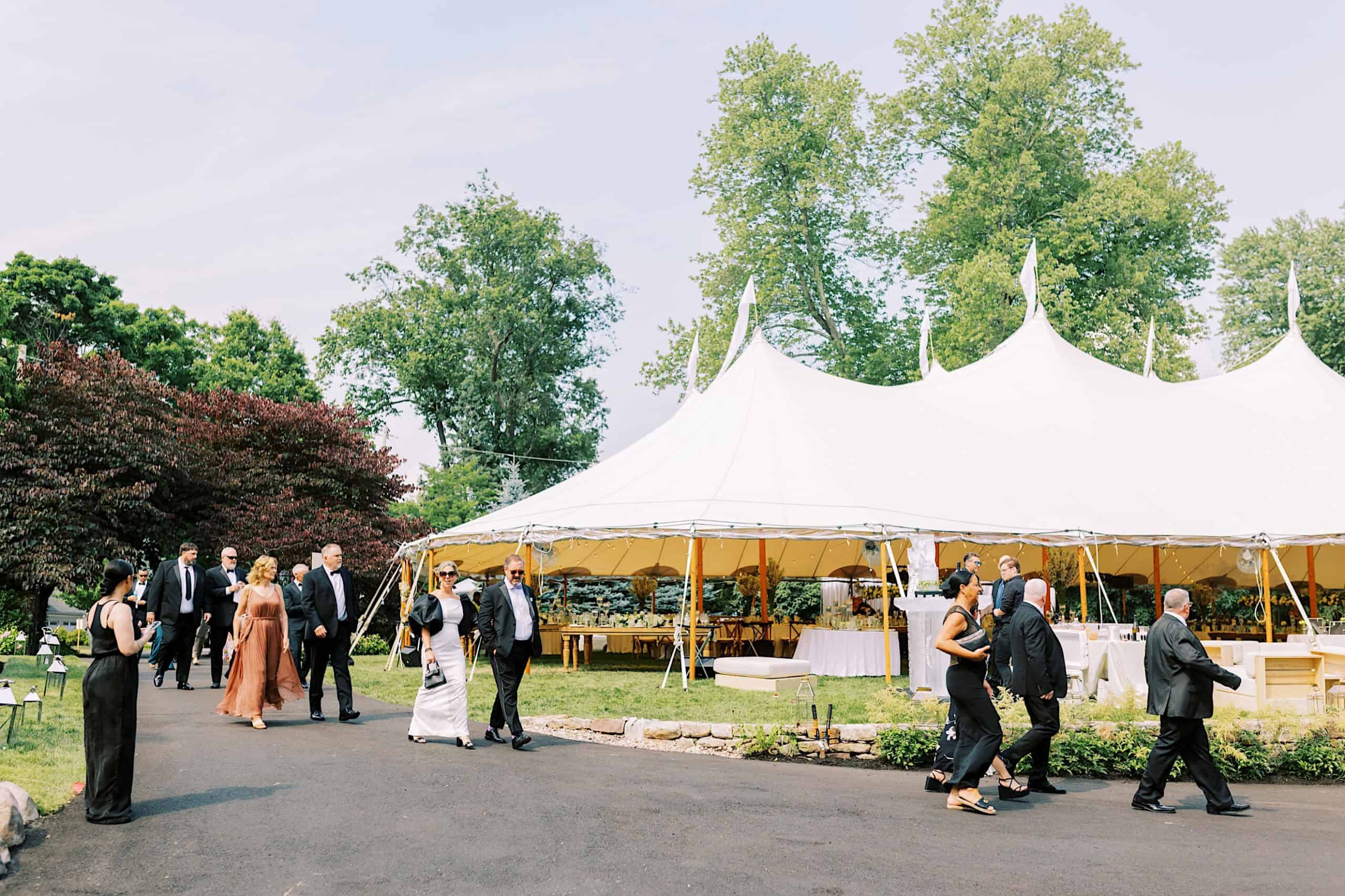 A group of people in formal attire walk toward a large white event tent set up outdoors on a sunny day, ready to celebrate a romantic backyard wedding in NJ.