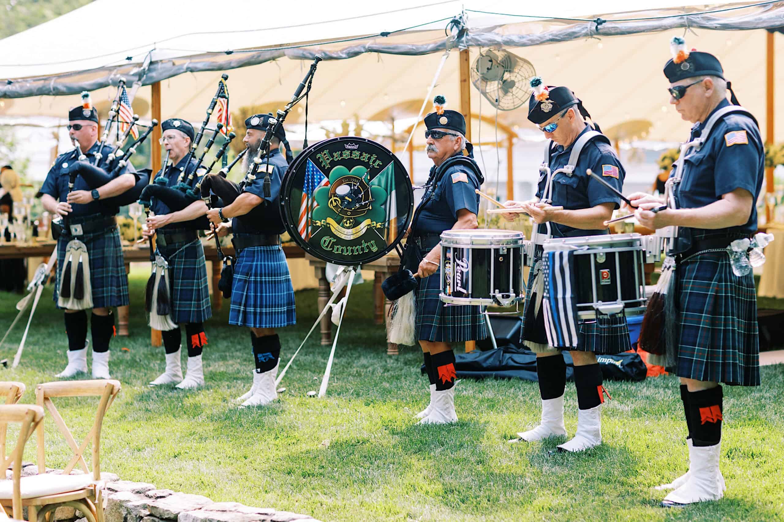 A group of uniformed bagpipers and drummers perform outdoors under a tent, with instruments and banners displaying “Emerald Society”—perfect entertainment for a romantic backyard wedding in NJ.