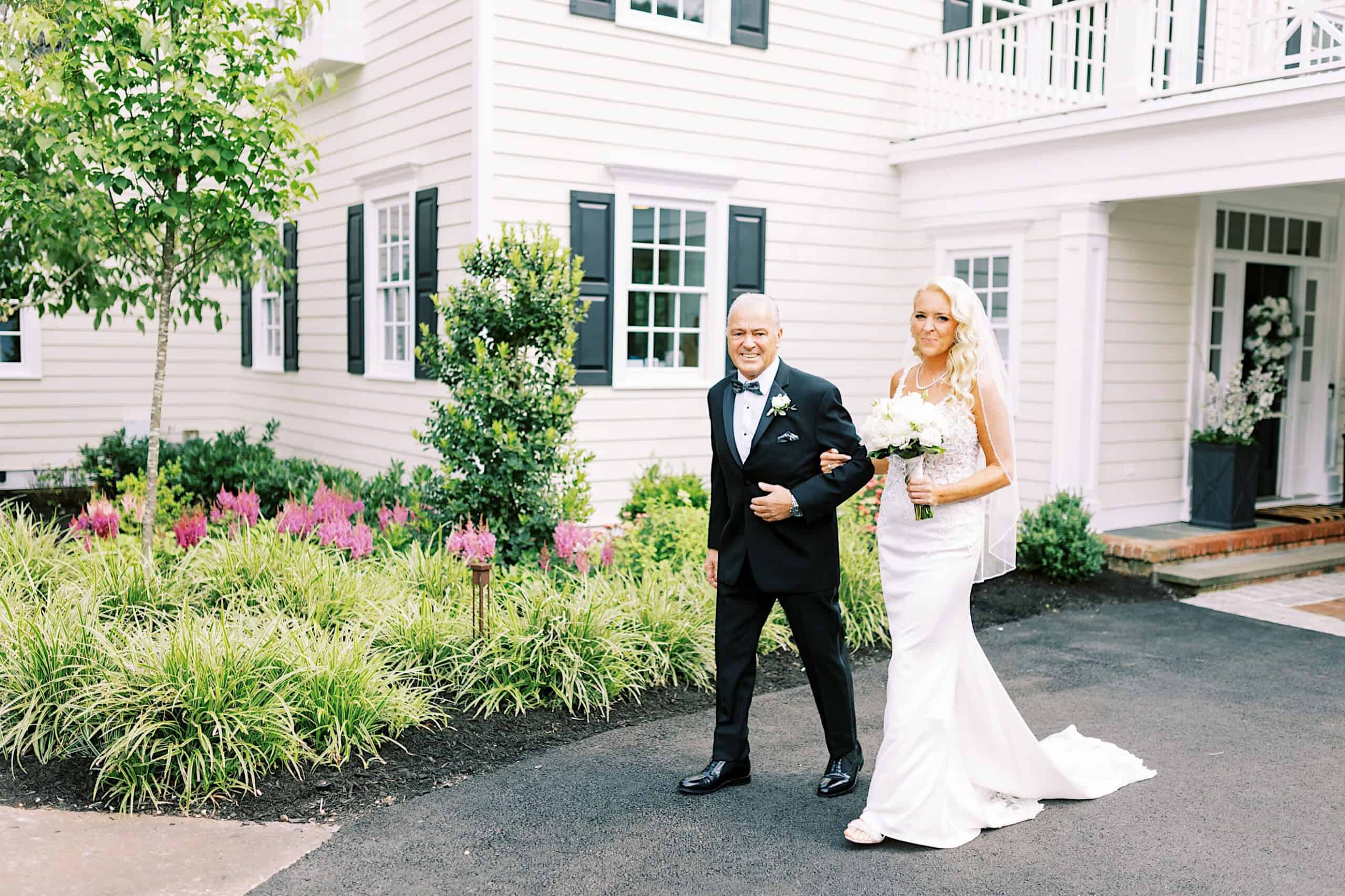A bride in a white dress holds a bouquet and walks arm-in-arm with an older man outside a white house with black shutters and landscaped greenery, capturing the charm of a romantic backyard wedding in NJ.