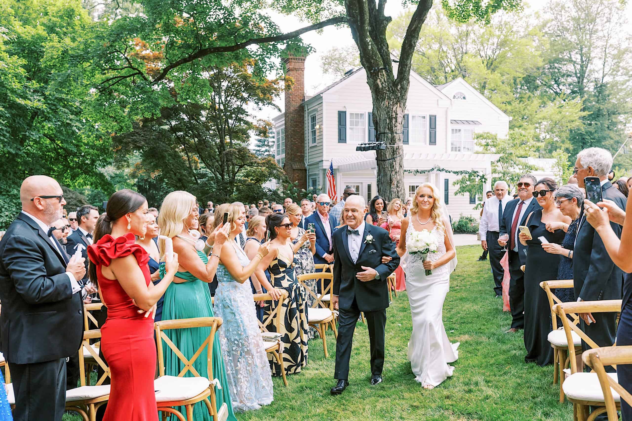 A bride in a white dress walks down the aisle with an older man, surrounded by guests seated outdoors at a romantic backyard wedding in NJ.