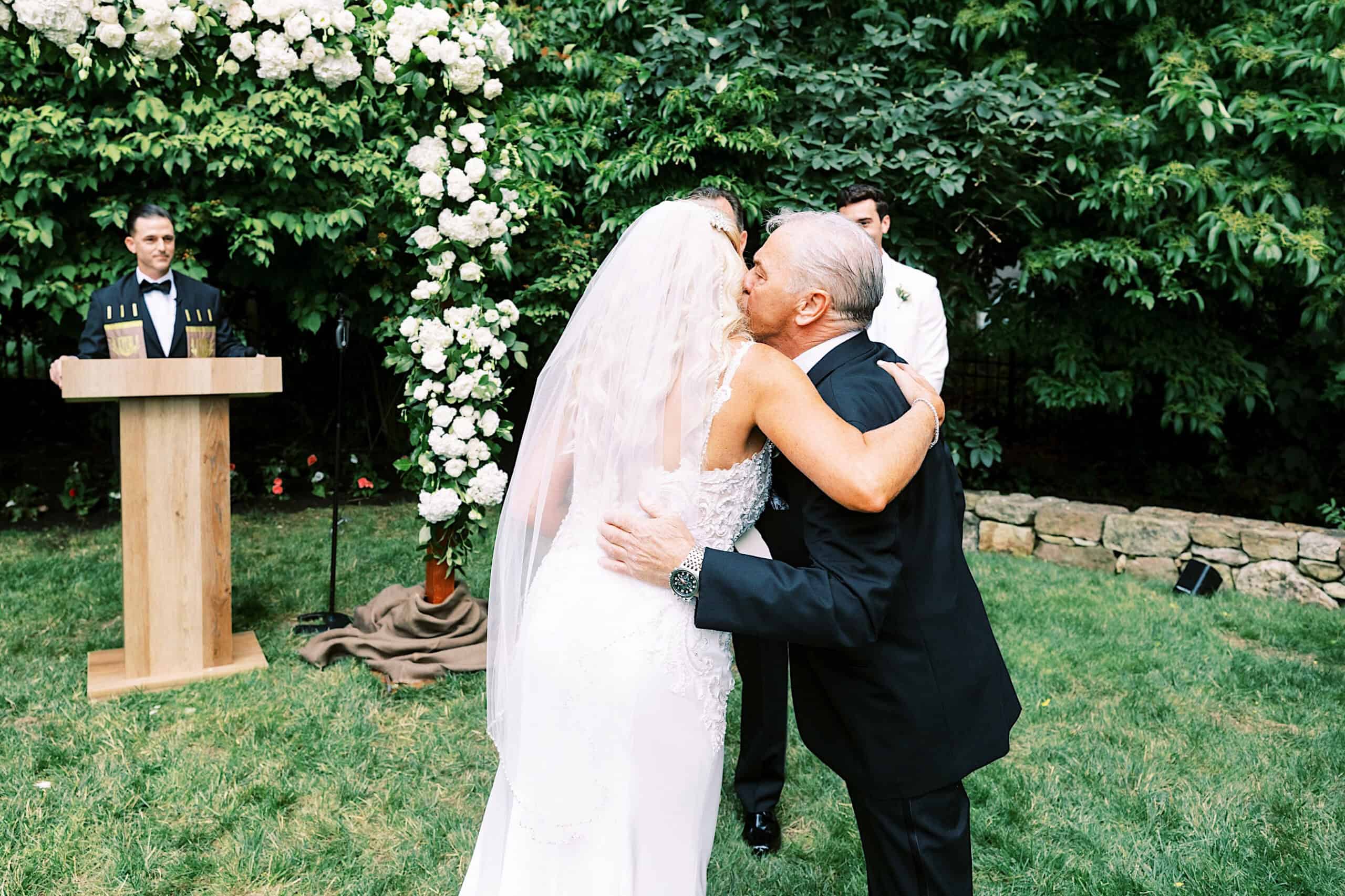 At a romantic backyard wedding in NJ, a bride in a white dress and veil hugs an older man in a suit, with an officiant and floral arch enhancing the heartfelt outdoor ceremony.