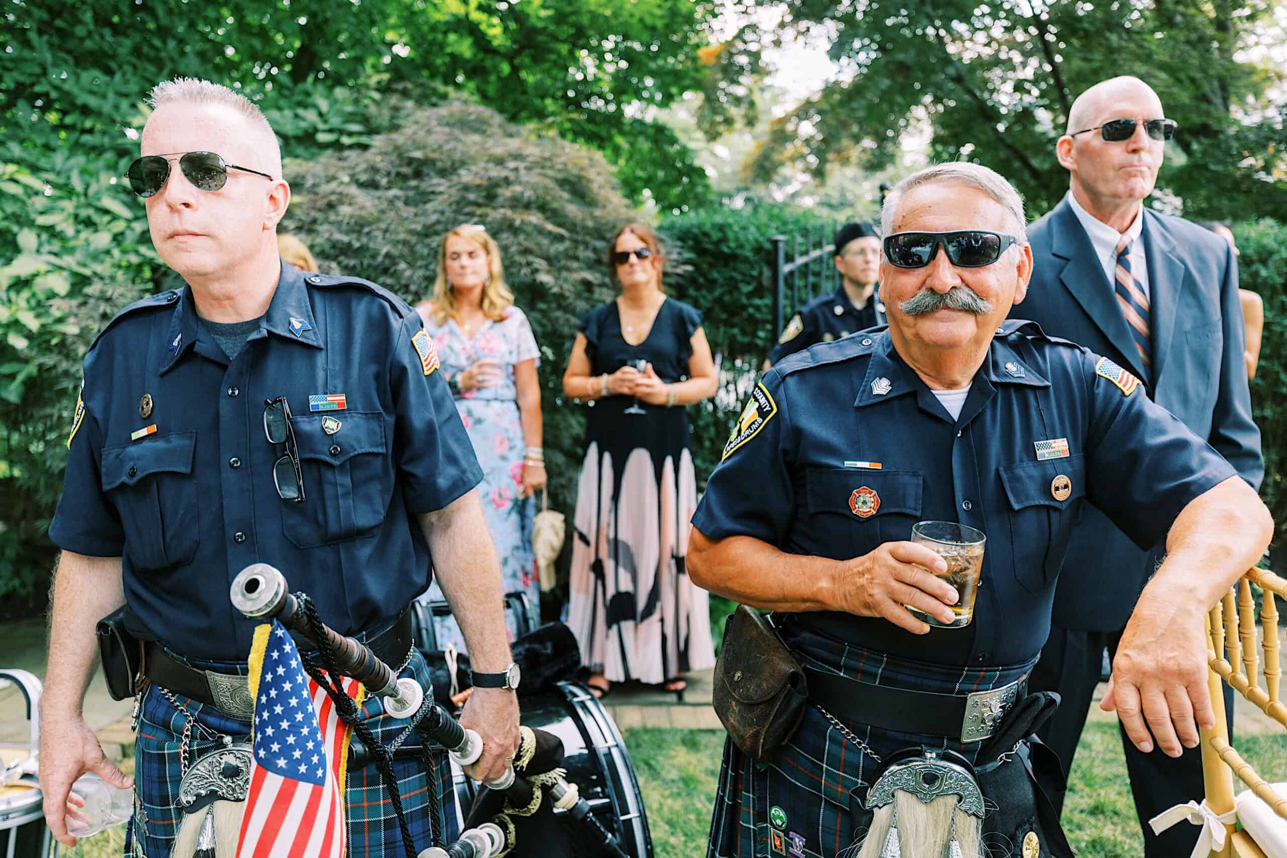 Two uniformed police officers in kilts stand outdoors with bagpipes at a romantic backyard wedding in NJ; one holds a drink while guests in formal attire gather in the background.