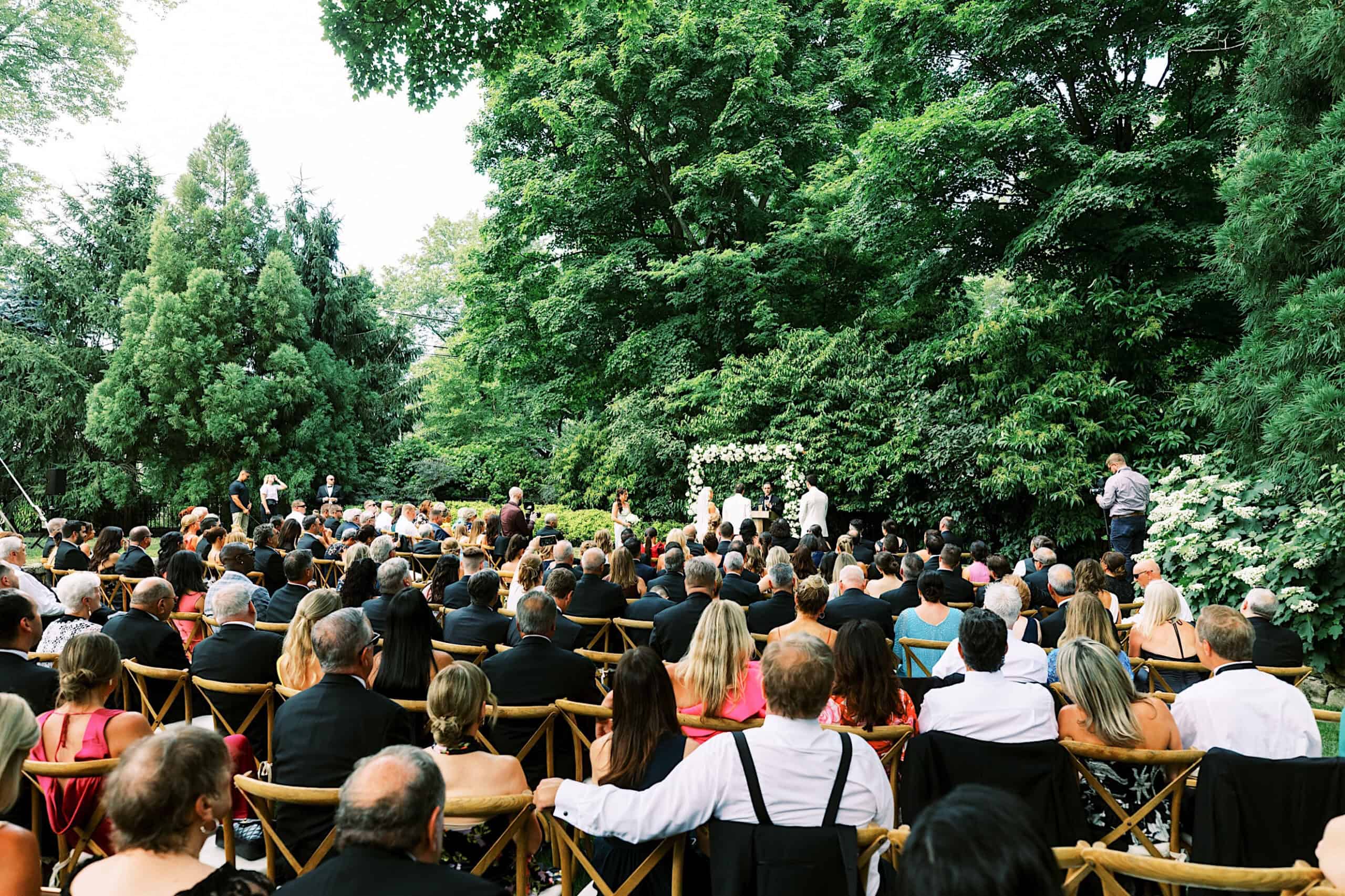 Romantic backyard wedding in NJ with seated guests facing a flower-adorned altar, surrounded by lush green trees and foliage for an unforgettable outdoor ceremony.