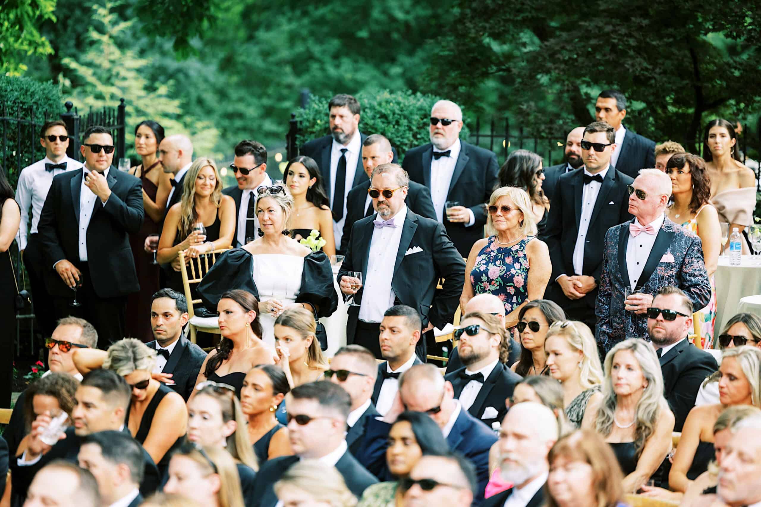A large group of people dressed in formal attire, seated and standing outdoors, attentively watching a romantic backyard wedding in NJ.