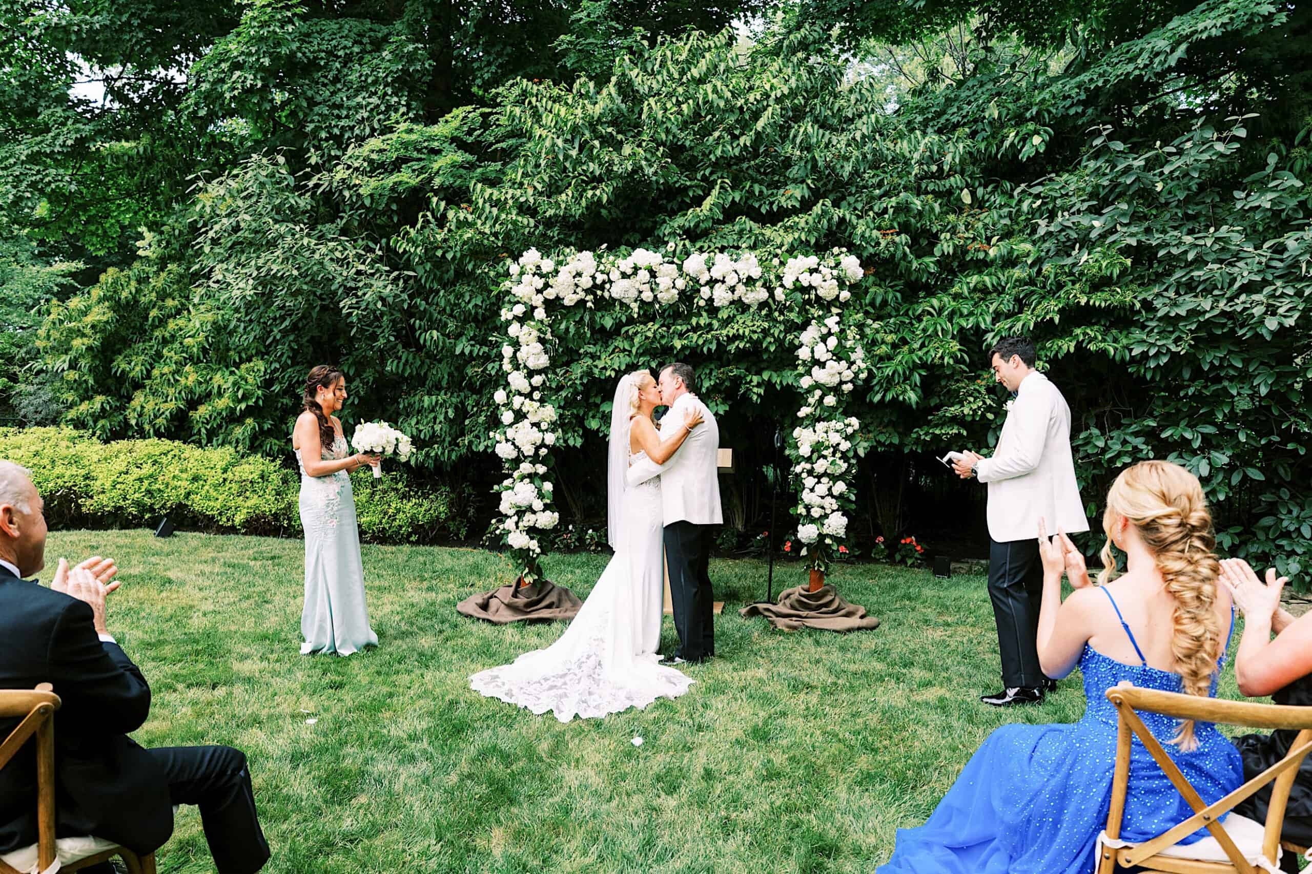 A bride and groom kiss under a floral arch at their romantic backyard wedding in NJ, surrounded by a small group of people clapping.