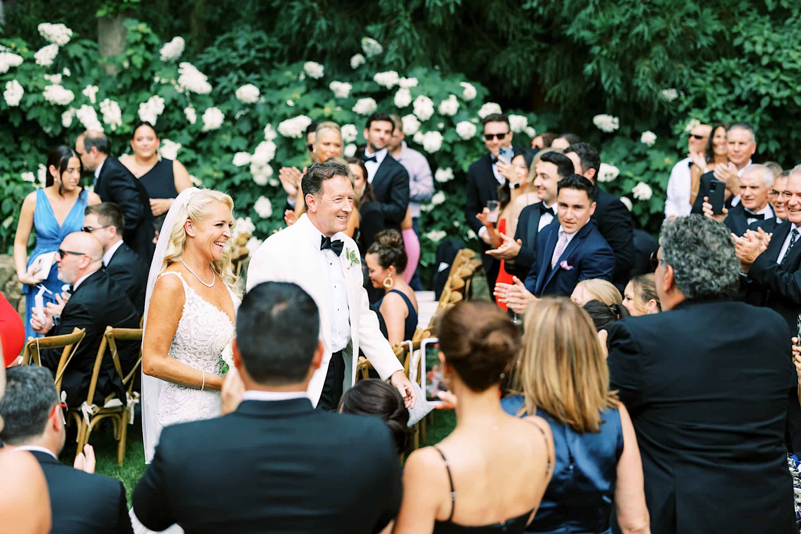 A bride and groom walk down an outdoor aisle at their romantic backyard wedding in NJ, smiling as guests in formal attire applaud and watch them amid greenery and white flowers.