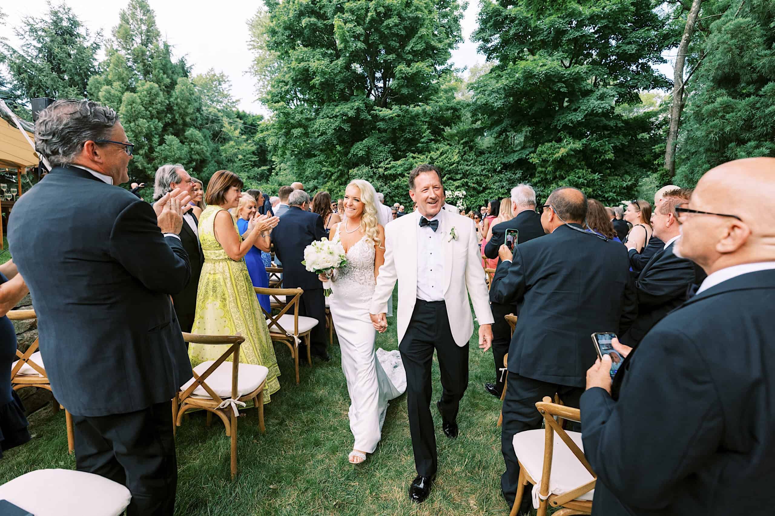 At a romantic backyard wedding in NJ, a bride and groom walk down an outdoor aisle smiling, surrounded by guests who are standing, clapping, and taking photos.