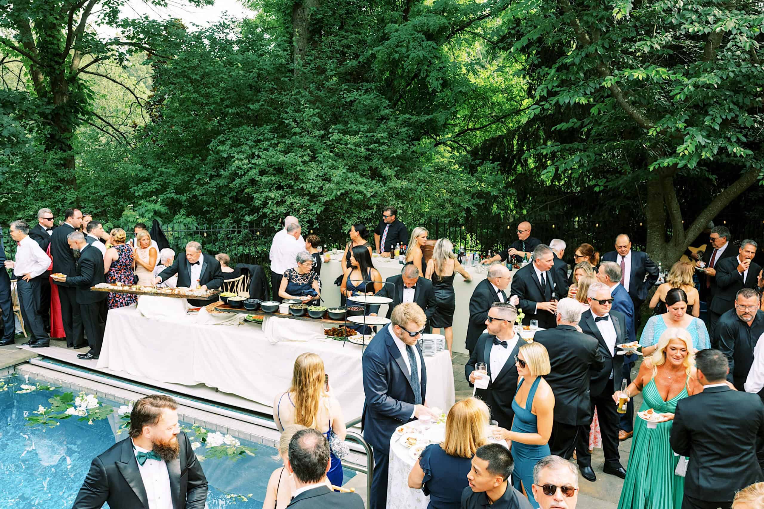 A large group of people dressed in formal attire gather around buffet tables at an outdoor poolside event, surrounded by trees, celebrating a romantic backyard wedding in NJ.