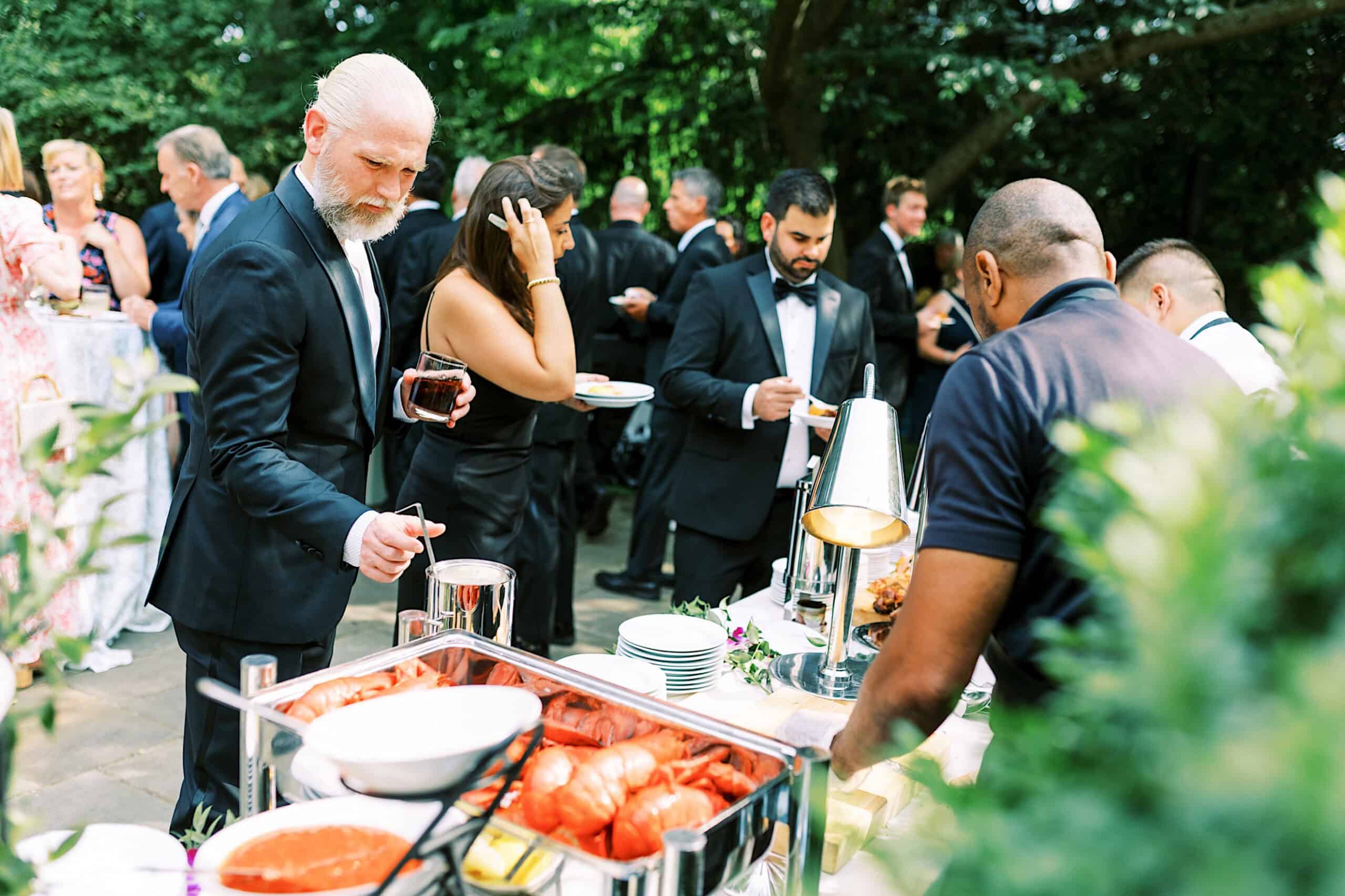 Guests in formal attire serve themselves food from a buffet table at a romantic backyard wedding in NJ, with trees and greenery in the background.