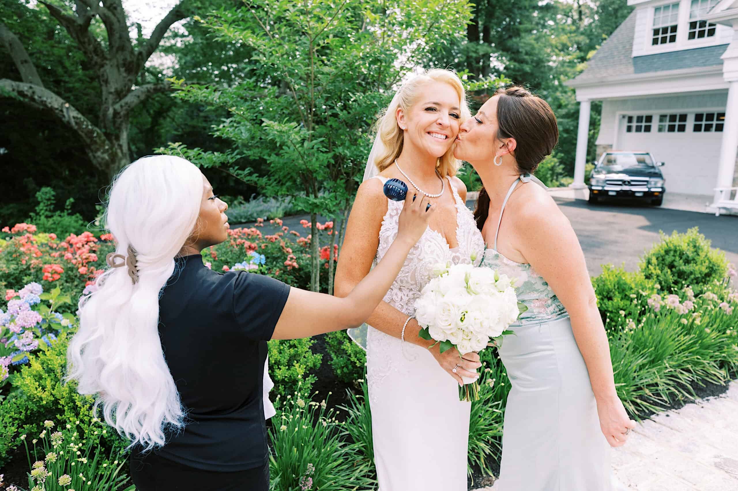 A bride smiles while holding a bouquet as another woman kisses her cheek during a romantic backyard wedding in NJ; nearby, a person with white hair adjusts her makeup outdoors near a house and garden.
