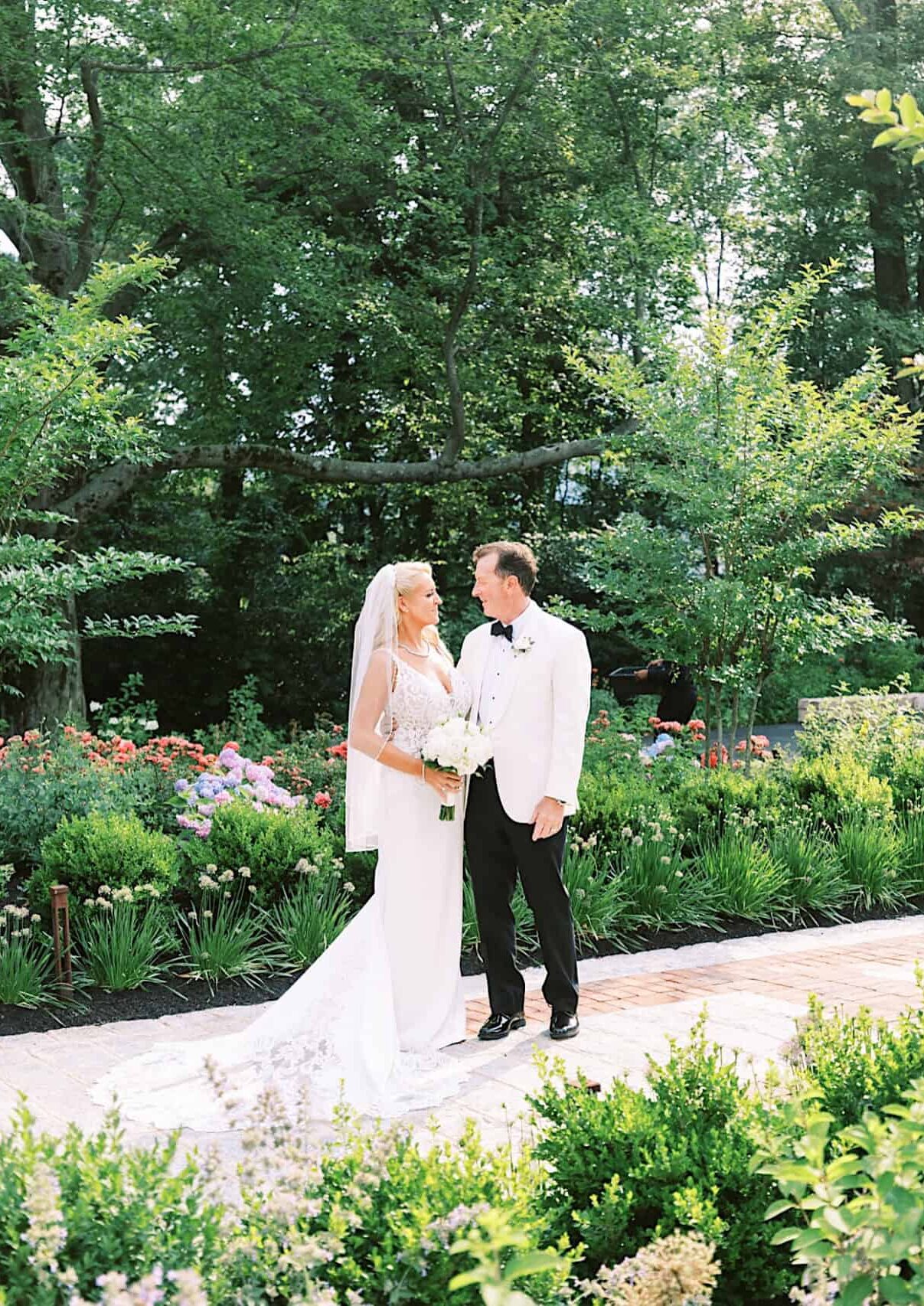 A bride in a white dress and a groom in a white jacket stand together on a garden path, surrounded by greenery and flowers during their romantic backyard wedding in NJ on a sunny day.