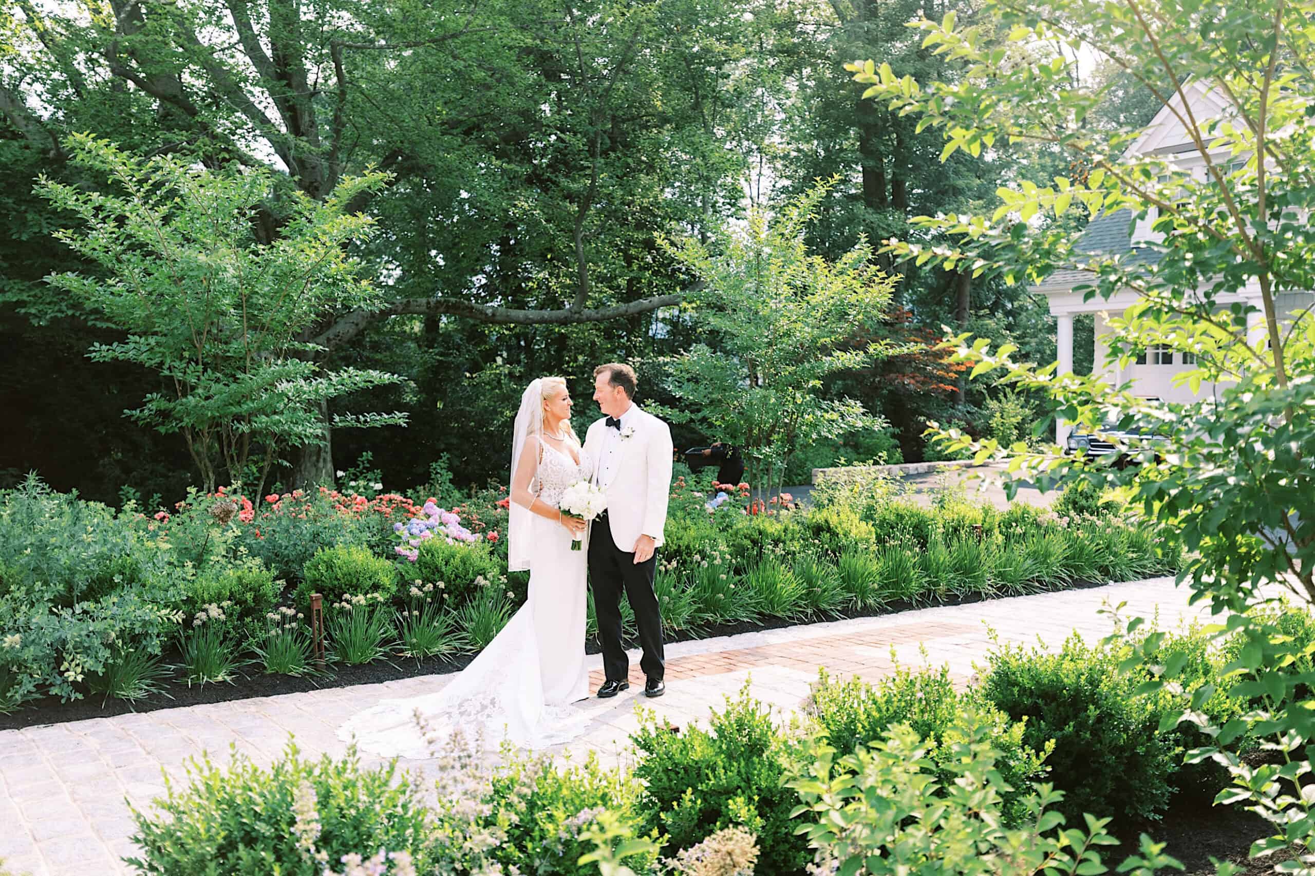 A bride in a white dress and a groom in a white jacket stand together on a garden path, surrounded by greenery and flowers during their romantic backyard wedding in NJ on a sunny day.