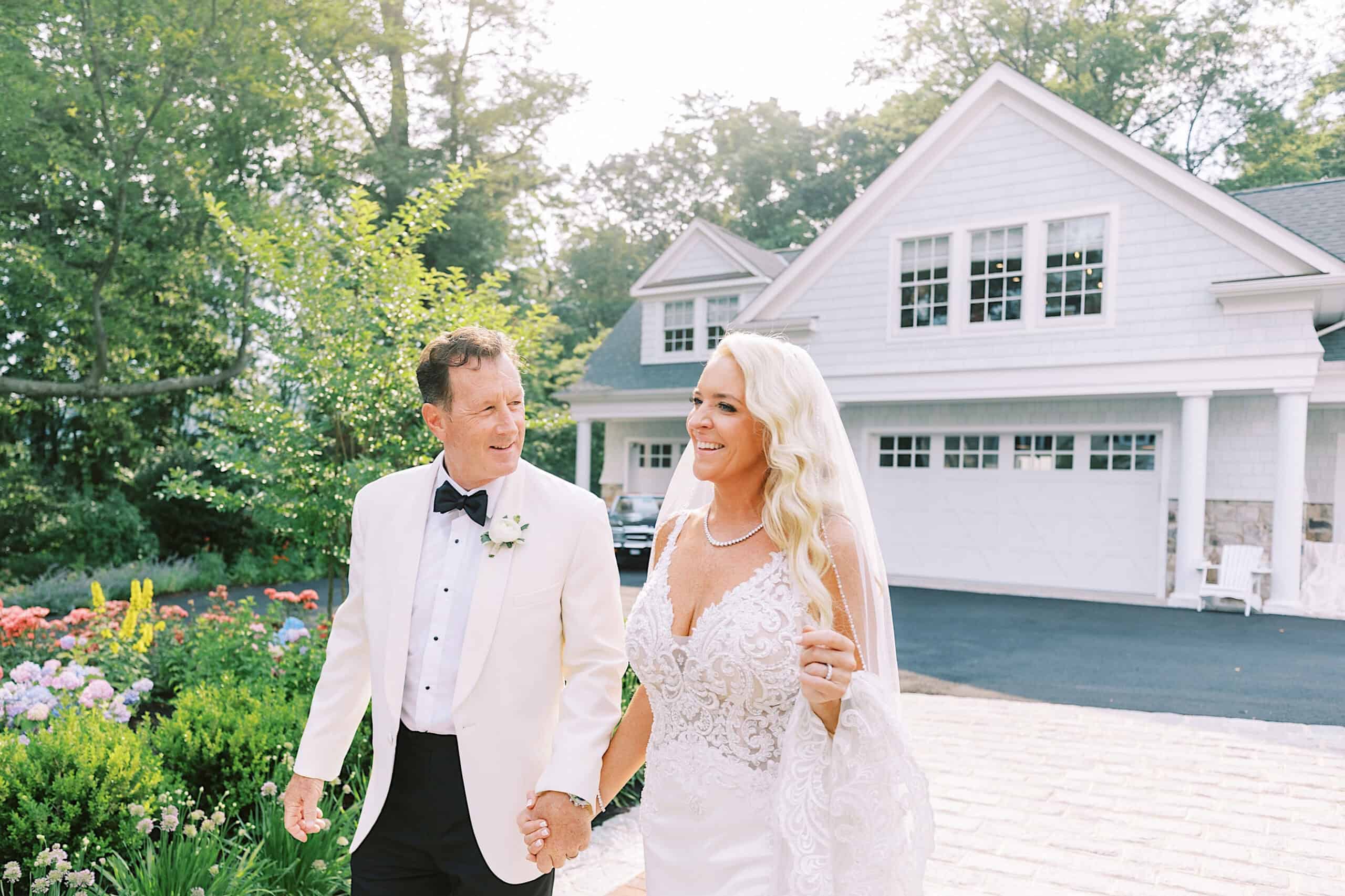 A man in a white tuxedo and a woman in a white wedding dress walk hand in hand outside a house, smiling at each other during their romantic backyard wedding in NJ.