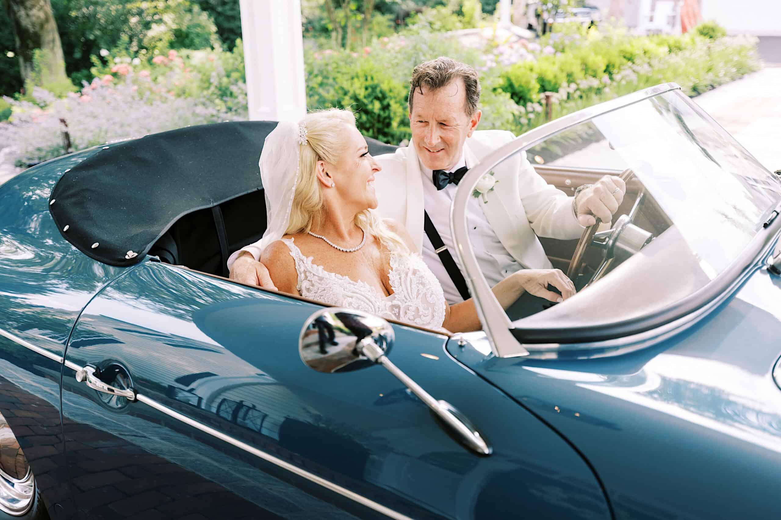 A bride and groom in wedding attire sit together in a vintage blue convertible car, smiling at each other after their romantic backyard wedding in NJ.