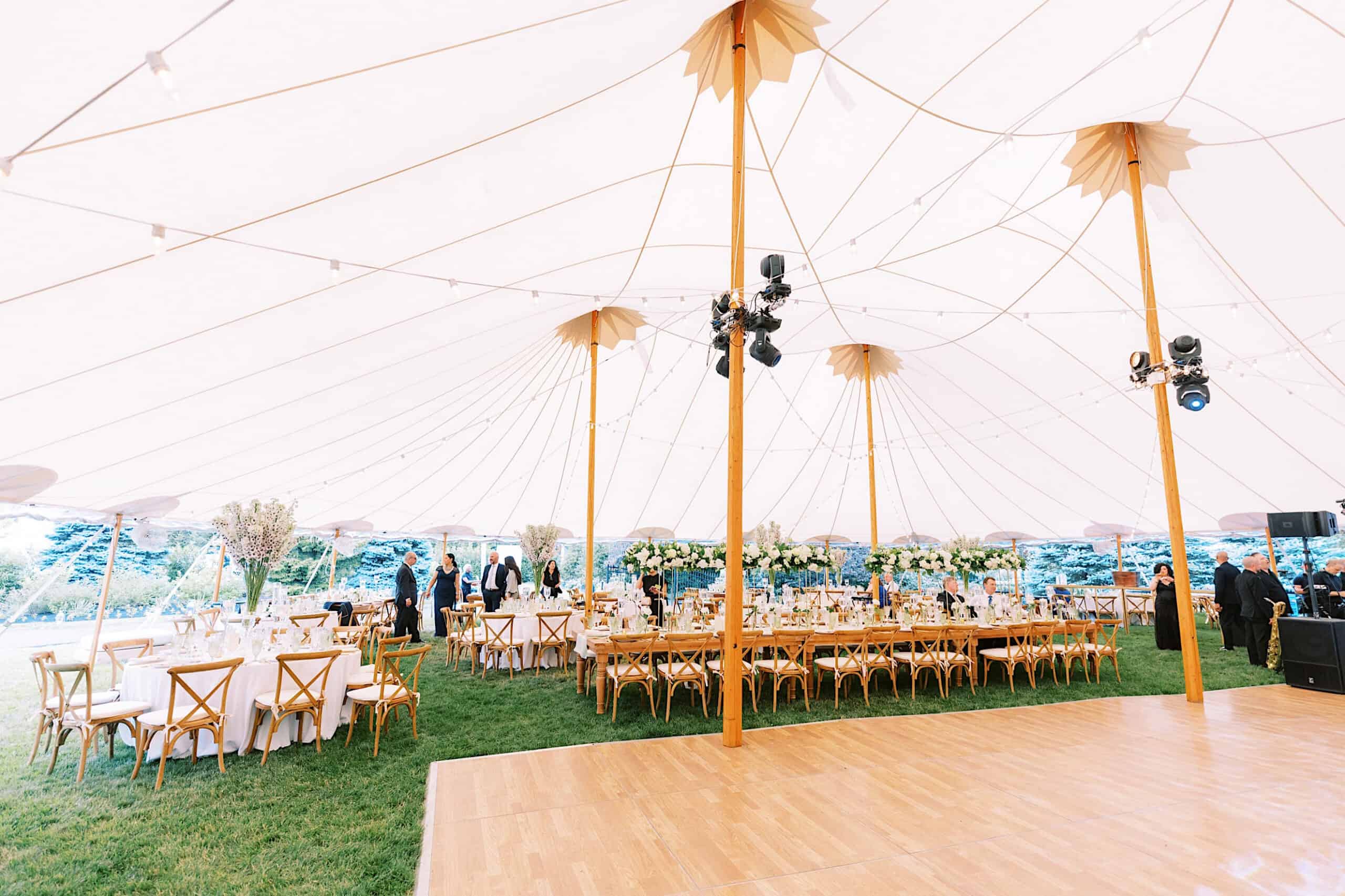 Large outdoor tent with wooden poles, tables for dining, wooden chairs, string lights, and a dance floor on grass—ideal for a romantic backyard wedding in NJ. People are setting up inside, creating an inviting celebration space.