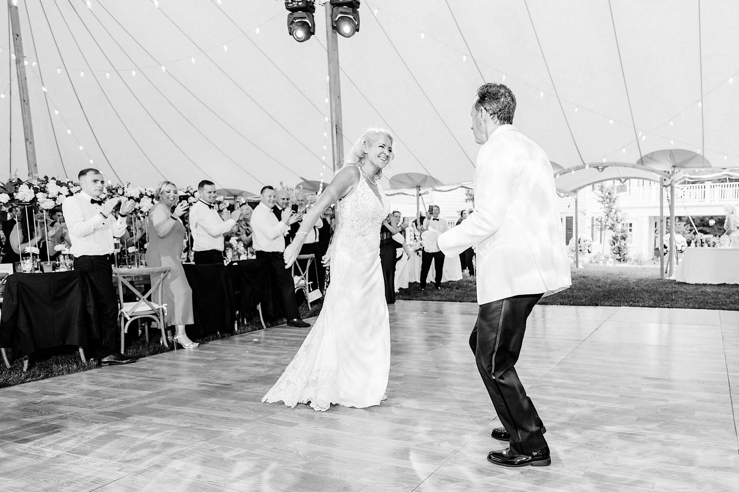 A bride and groom share a dance on a wooden floor under a tent as guests watch and applaud during their romantic backyard wedding in NJ.