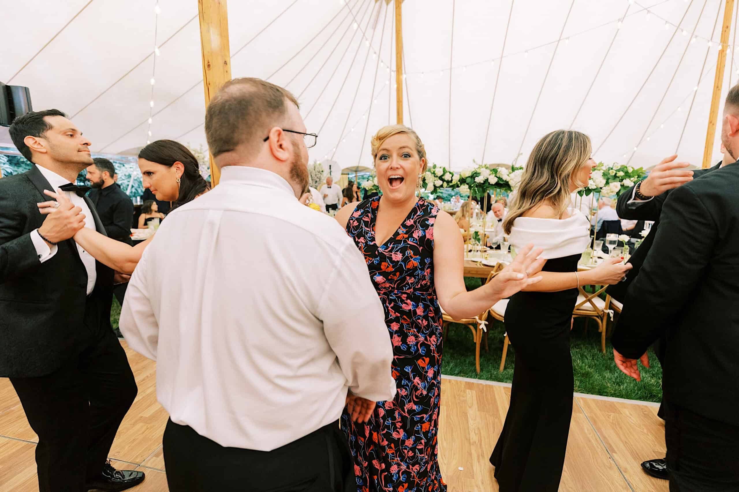 A group of people in formal attire dance and socialize under a large tent decorated with string lights and flower arrangements at a romantic backyard wedding in NJ.