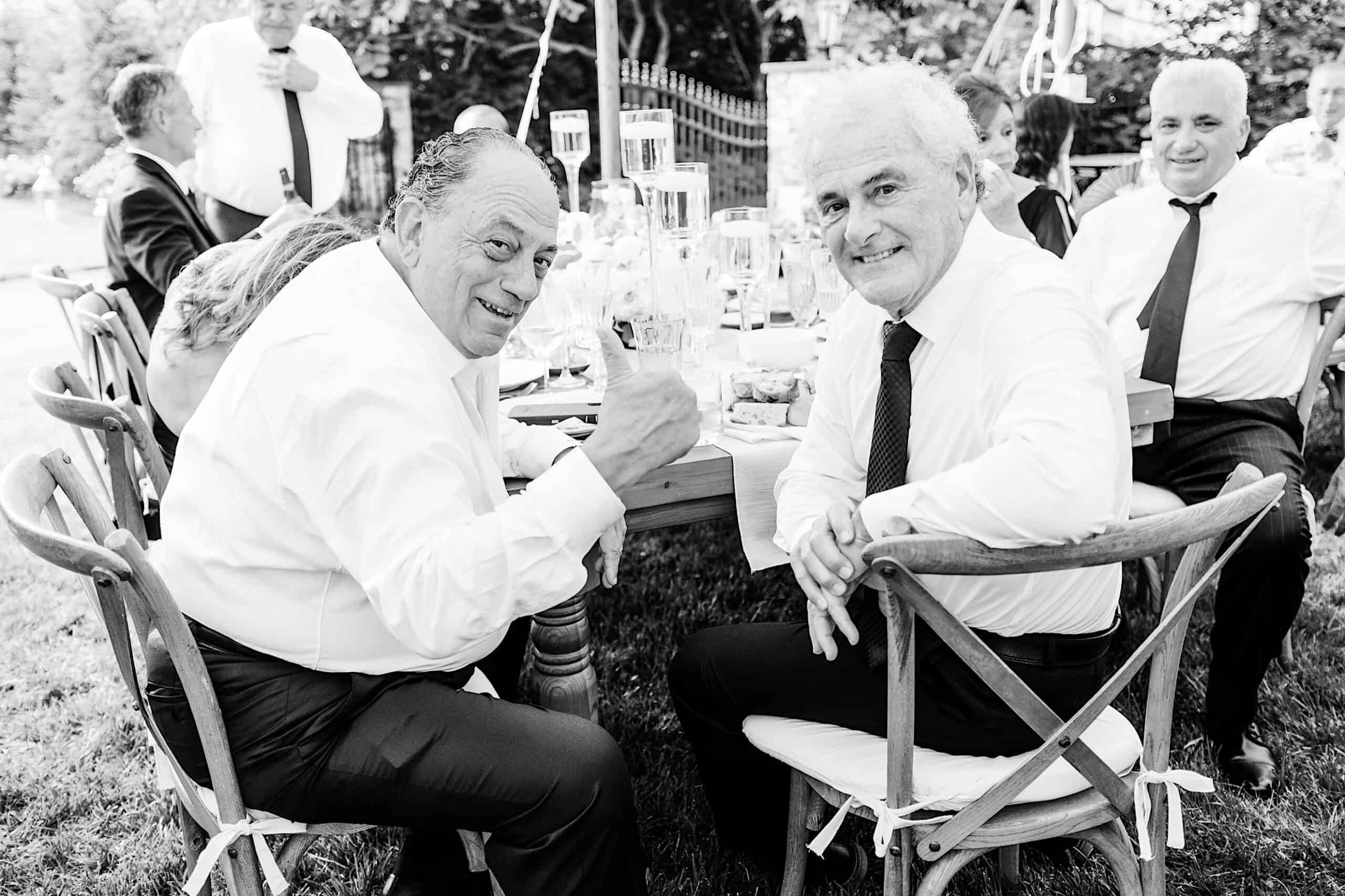 Two older men in dress shirts and ties sit at an outdoor table, smiling at the camera with other guests dining around them at a romantic backyard wedding in NJ.