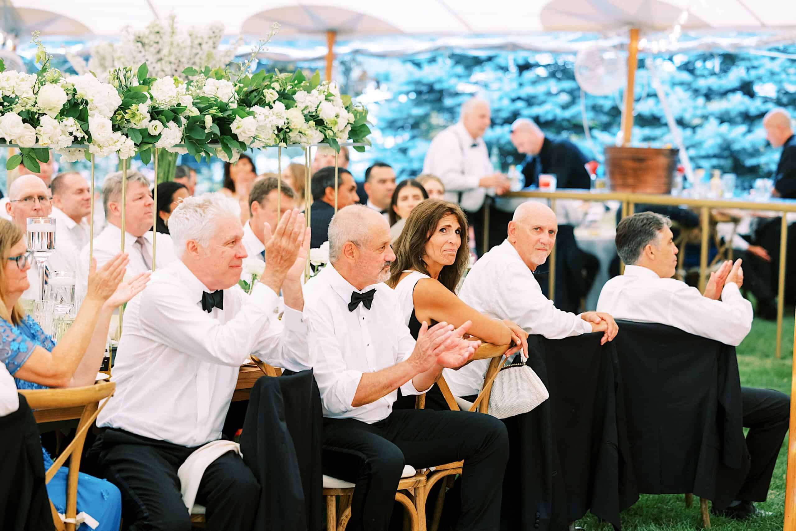 A group of people dressed formally sit at tables under a tent, applauding and watching an event, with floral centerpieces visible on the tables at a romantic backyard wedding in NJ.