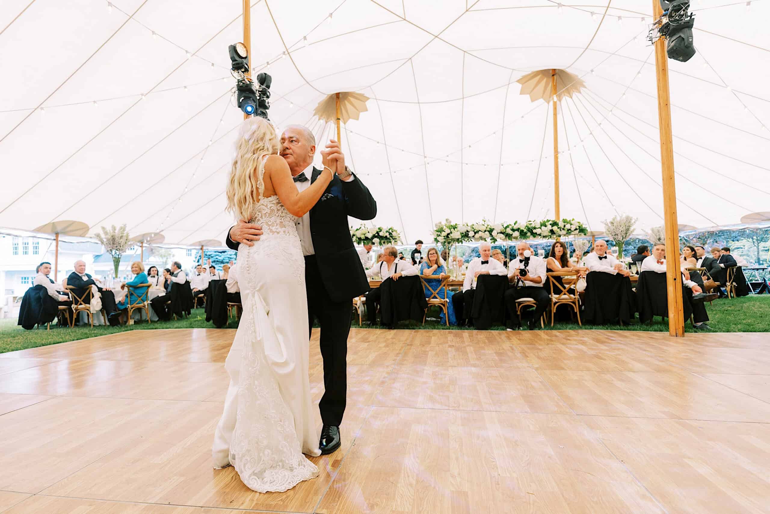 At a romantic backyard wedding in NJ, a bride in a white dress dances with an older man on a wooden floor under a large white tent, as guests seated at tables watch.