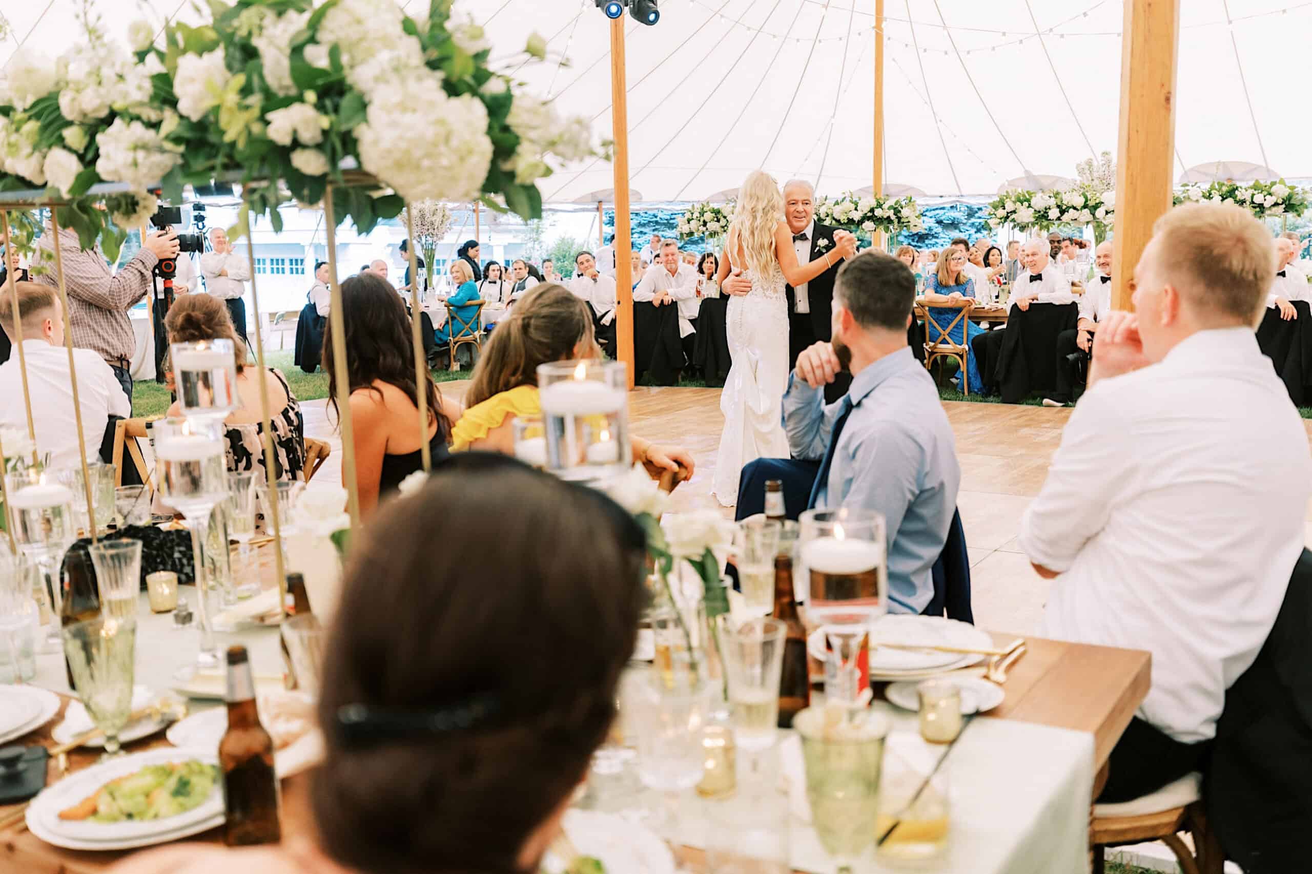 A woman in a white dress stands and speaks to seated guests at a romantic backyard wedding in NJ, held in a decorated tent with floral centerpieces and candlelit tables.