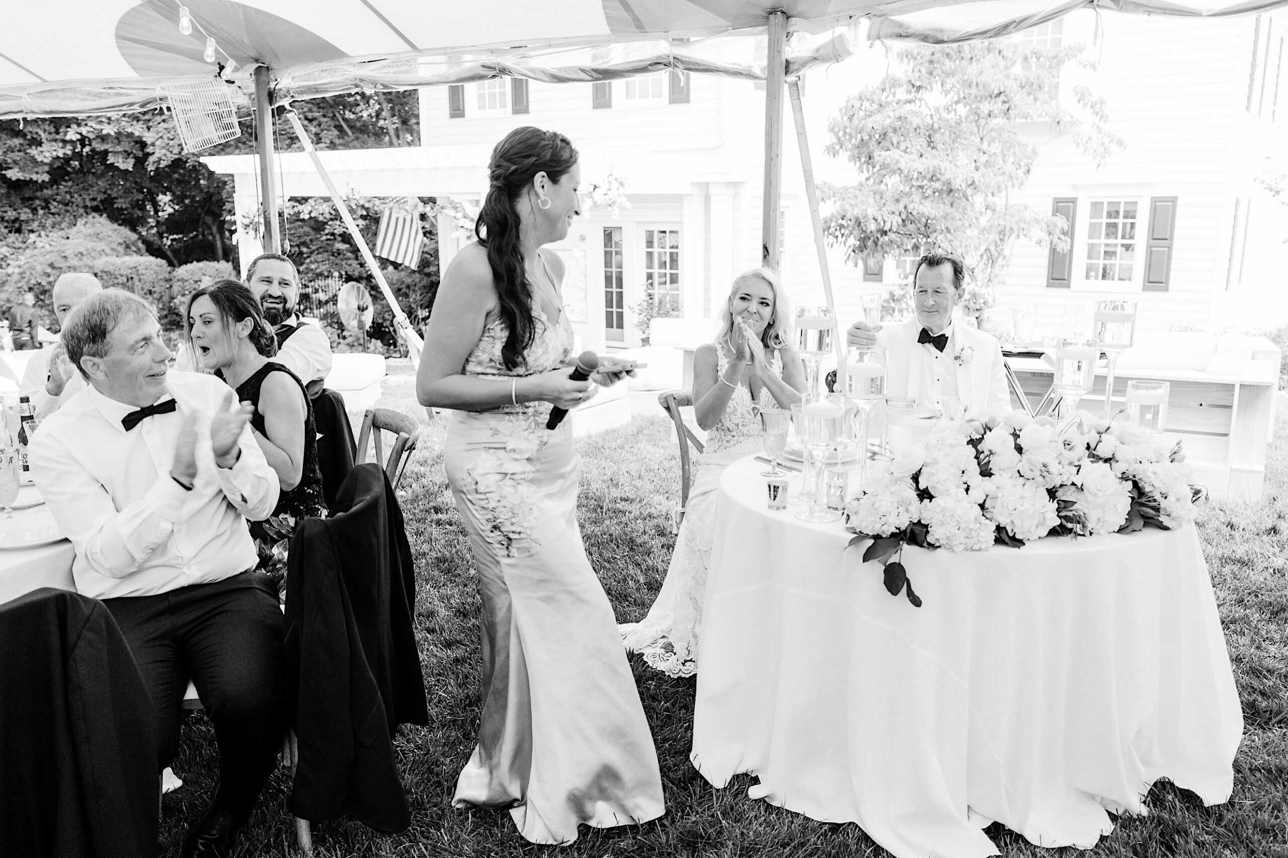 A woman in a formal dress stands and speaks at a romantic backyard wedding in NJ while guests and the seated couple listen and clap.