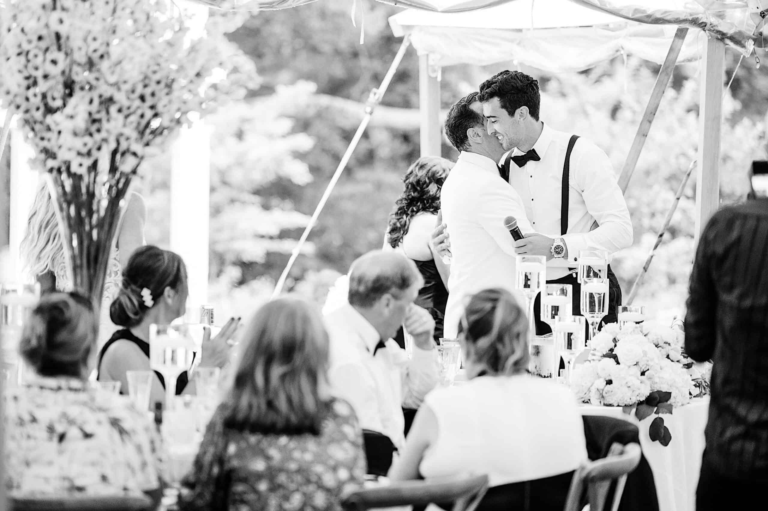 Two men in formal wear share a kiss under a canopy at their romantic backyard wedding in NJ, while guests seated at tables look on.