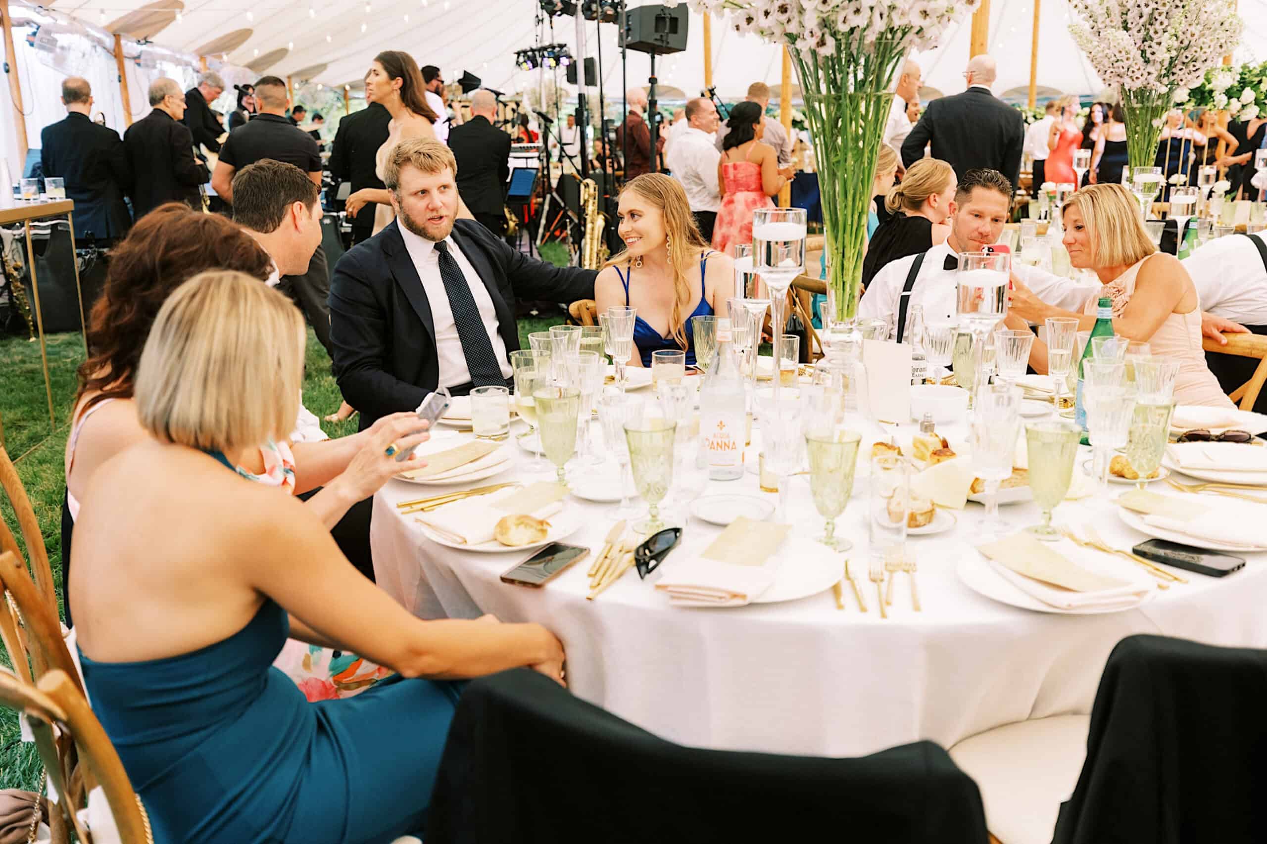 A group of people dressed in formal attire sit around a round table set for a meal at a large, elegant romantic backyard wedding in NJ inside a beautifully decorated tent.