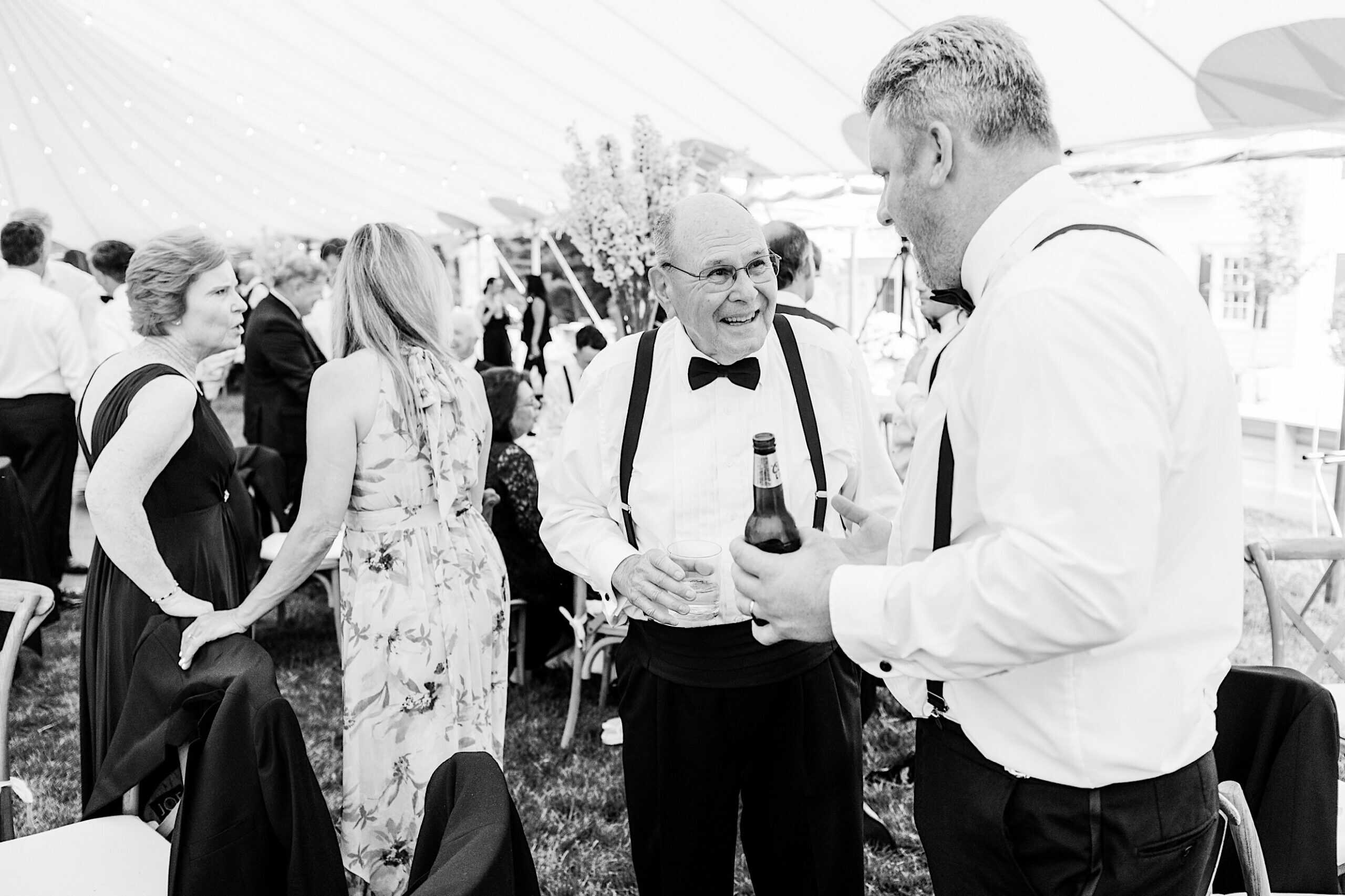 People in formal attire mingle under a tent at a romantic backyard wedding in NJ; one man holds a beer bottle while chatting with an older gentleman sporting suspenders and a bow tie.