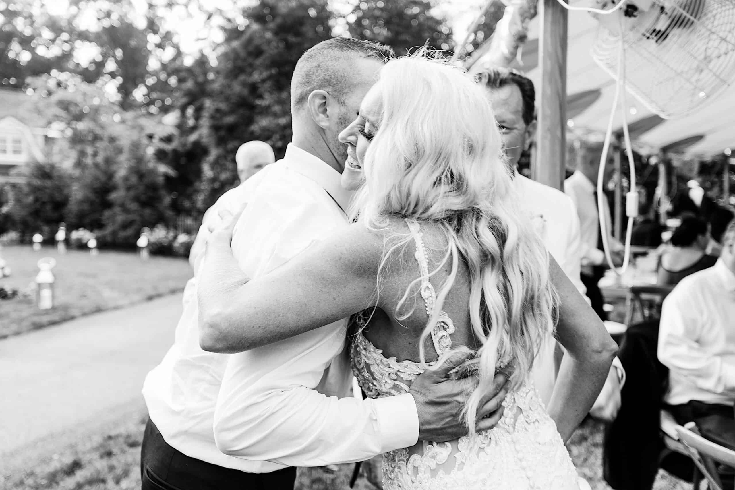 A bride and a man embrace and smile at their romantic backyard wedding in NJ, surrounded by guests under a tent at an outdoor reception.
