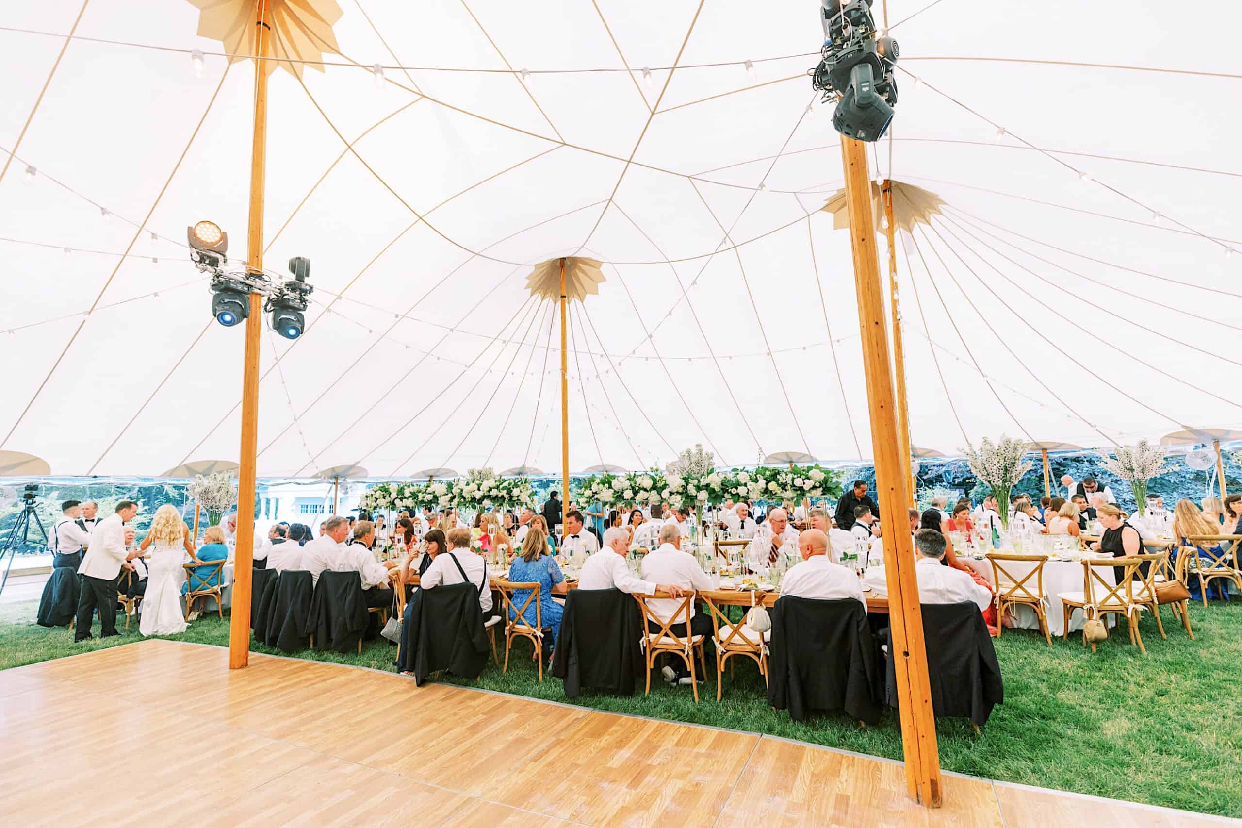 Guests seated at round tables under a large white tent at a romantic backyard wedding in NJ, with floral centerpieces and wooden chairs set on a grassy area.