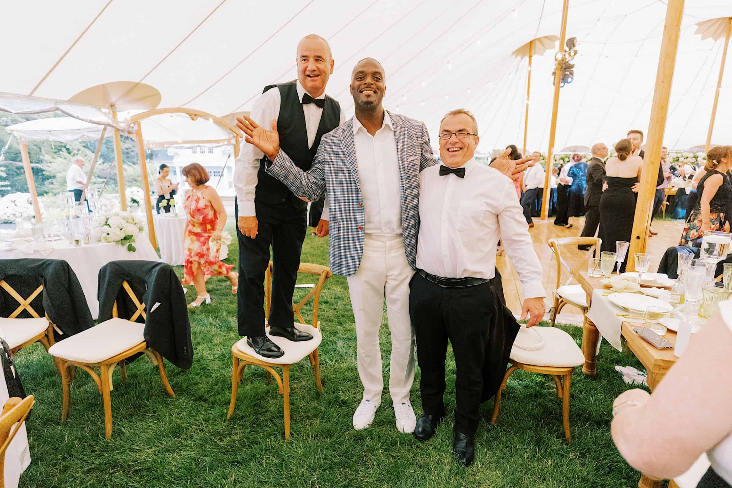 Three men pose and smile under a large event tent at a romantic backyard wedding in NJ; two stand on chairs, one stands on the ground, with tables, chairs, and other guests in the background.