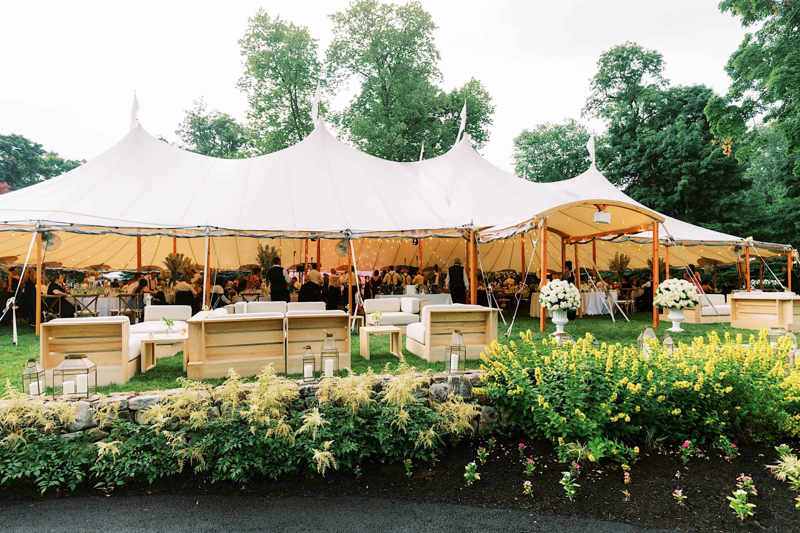 Large white outdoor event tent set for a romantic backyard wedding in NJ, with guests seated inside, wooden sofas and chairs on the lawn, surrounded by green trees and landscaped flower beds.