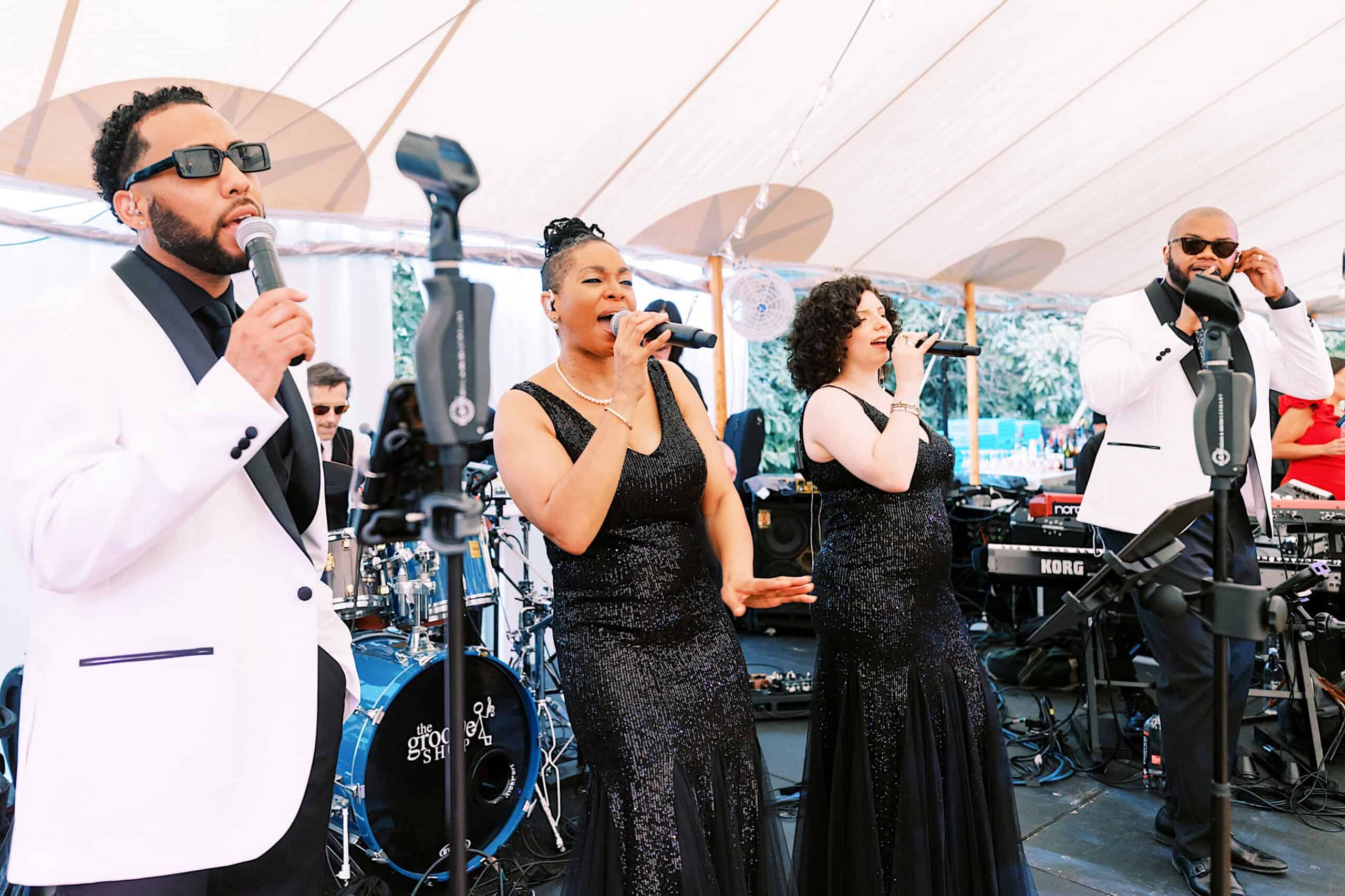Four musicians in formal attire perform on an outdoor stage at a romantic backyard wedding in NJ. Two men and two women sing into microphones, with a drum set and keyboard visible in the background.