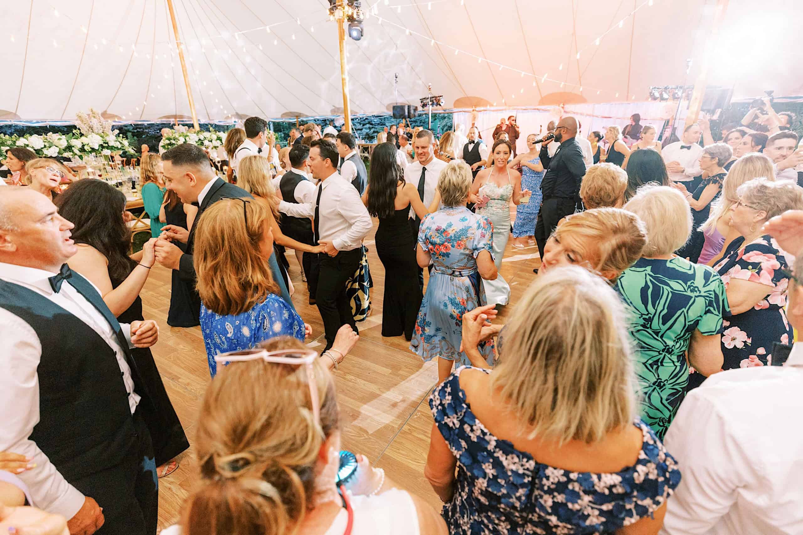A large group of people dressed in formal attire dance and socialize under a tent decorated with string lights at a romantic backyard wedding in NJ.
