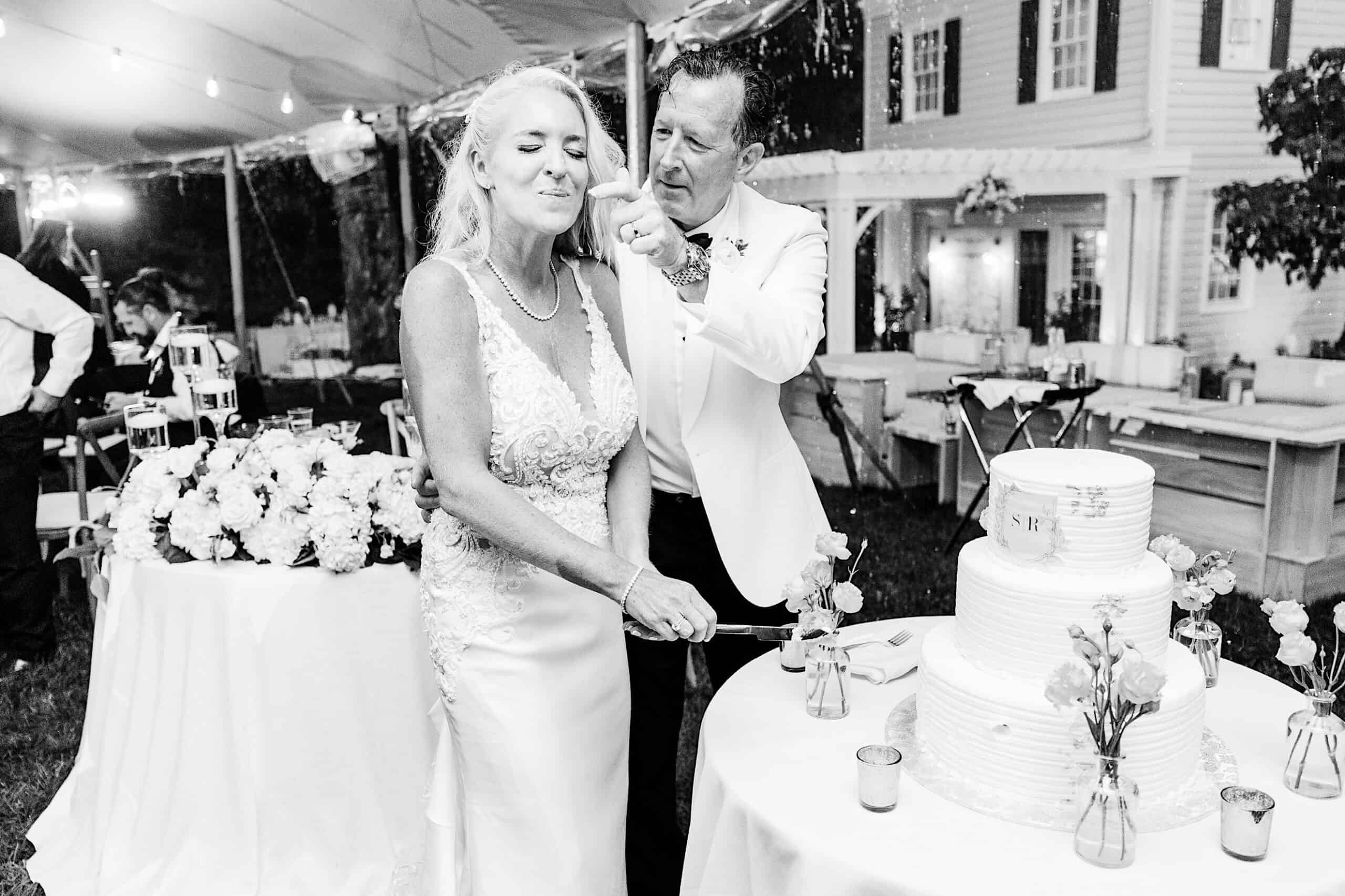 At a romantic backyard wedding in NJ, a bride and groom stand by the cake; the groom playfully feeds the bride as she smiles with eyes closed at their outdoor reception.