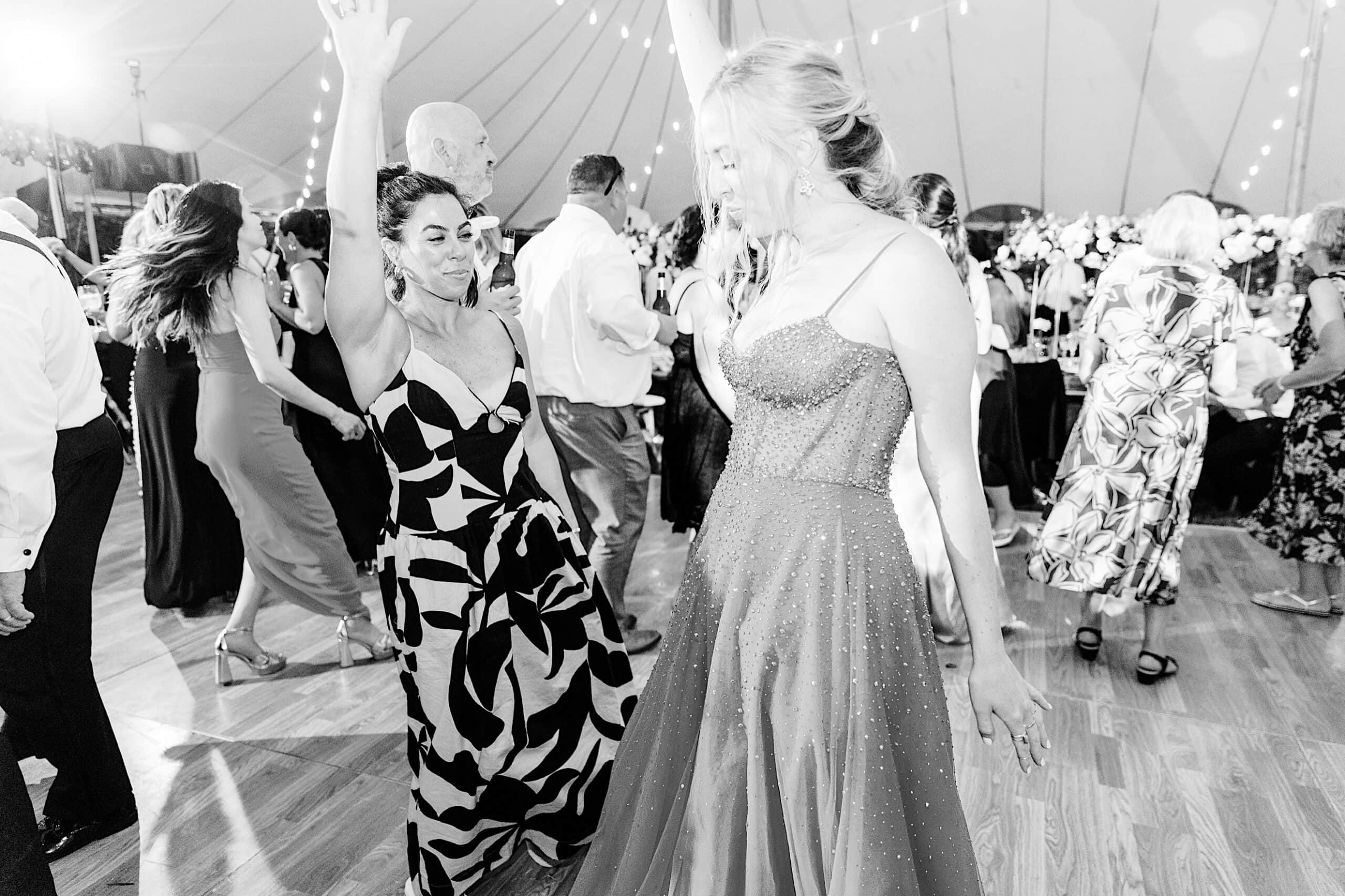 Two women in formal dresses dance on a crowded wooden floor under string lights at a romantic backyard wedding in NJ, surrounded by other guests.