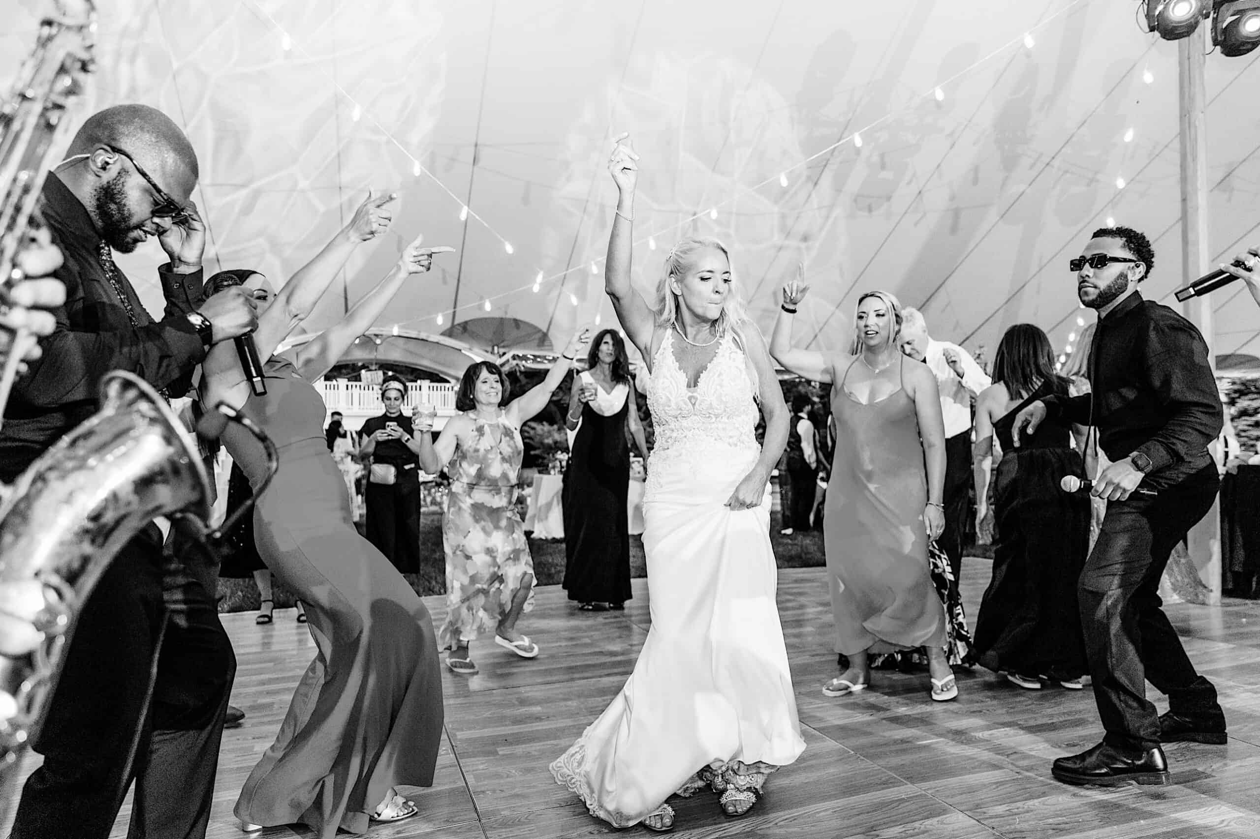 A bride dances energetically on a crowded dance floor with guests and musicians under a tent decorated with string lights at a romantic backyard wedding in NJ.