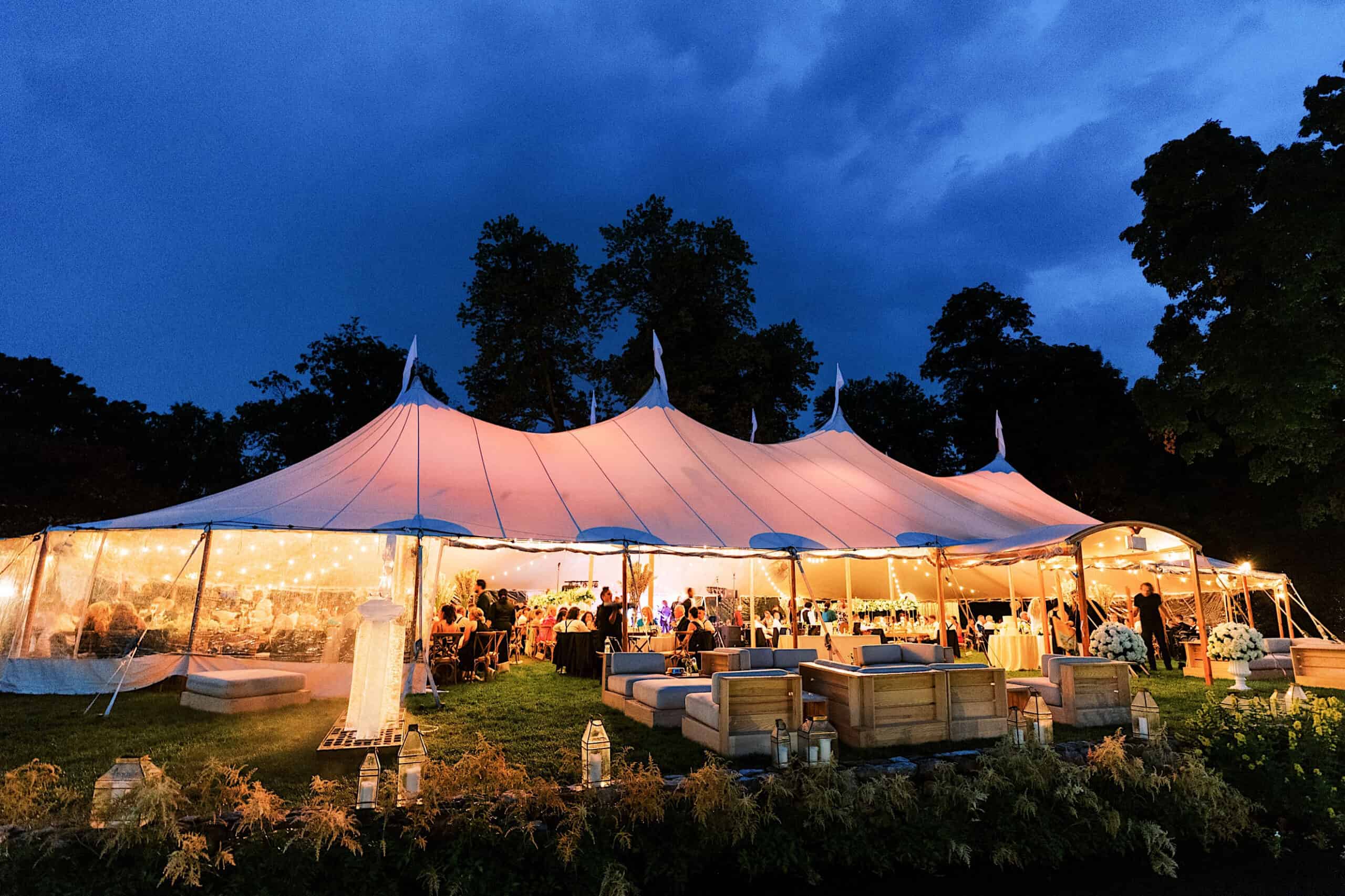 A large, white event tent is illuminated at dusk, with people gathered inside and around, creating the perfect setting for a romantic backyard wedding in NJ, surrounded by outdoor seating and trees in the background.