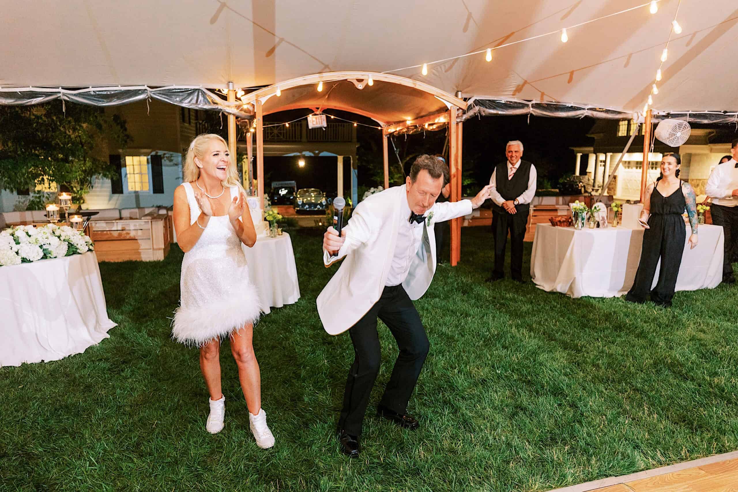 At a romantic backyard wedding in NJ, a man in a white suit energetically dances with a microphone while a woman in a short white dress and sneakers smiles under a tent at the joyful outdoor celebration.