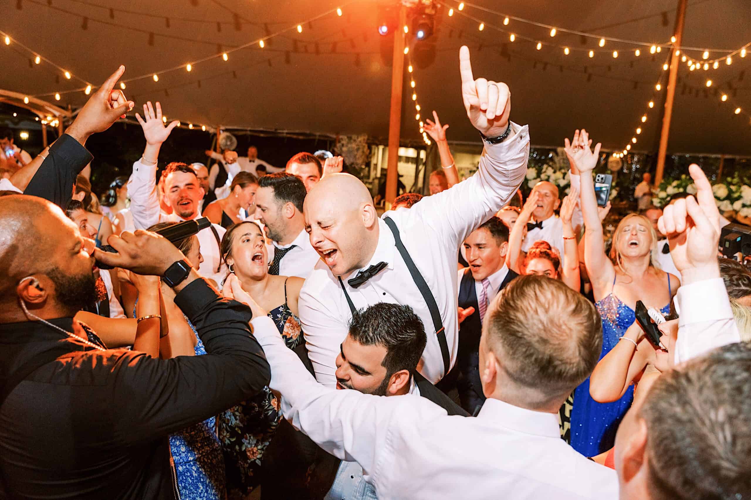 A large group of people dressed formally dance and celebrate energetically under string lights at a lively indoor event, reminiscent of a romantic backyard wedding in NJ.