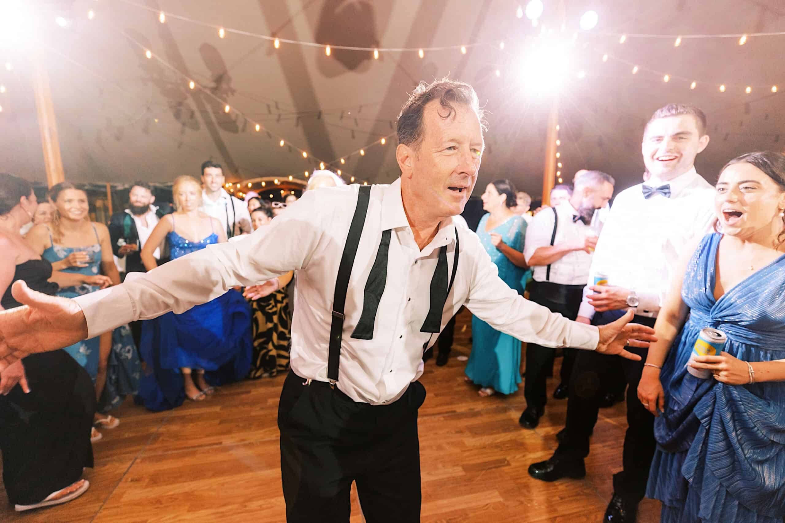 At a romantic backyard wedding in NJ, a man in formal attire with suspenders dances energetically on a wooden floor, surrounded by people in formal wear under string lights.