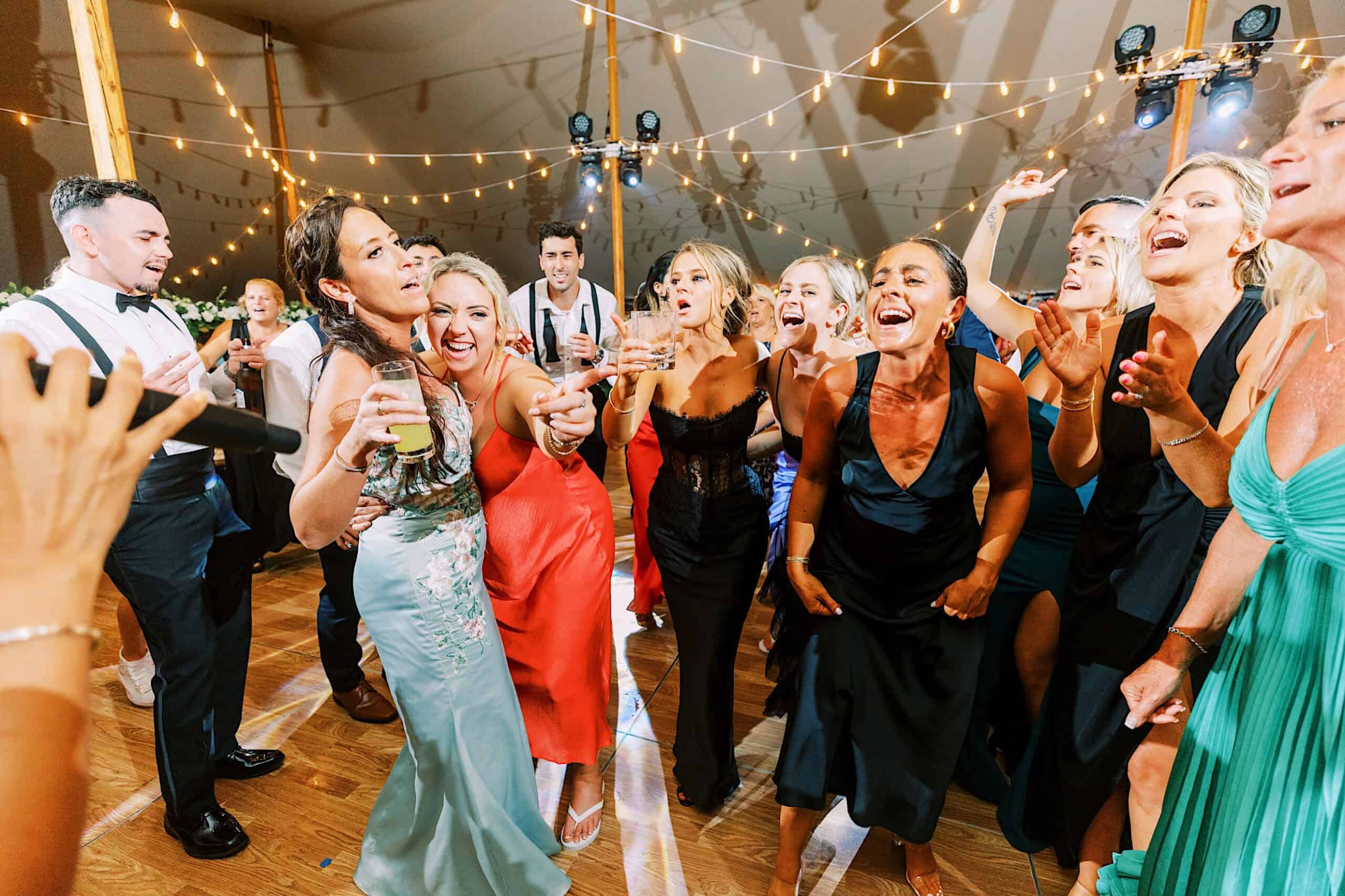A group of people in formal attire sing and dance together on a wooden floor under string lights at a lively romantic backyard wedding in NJ.