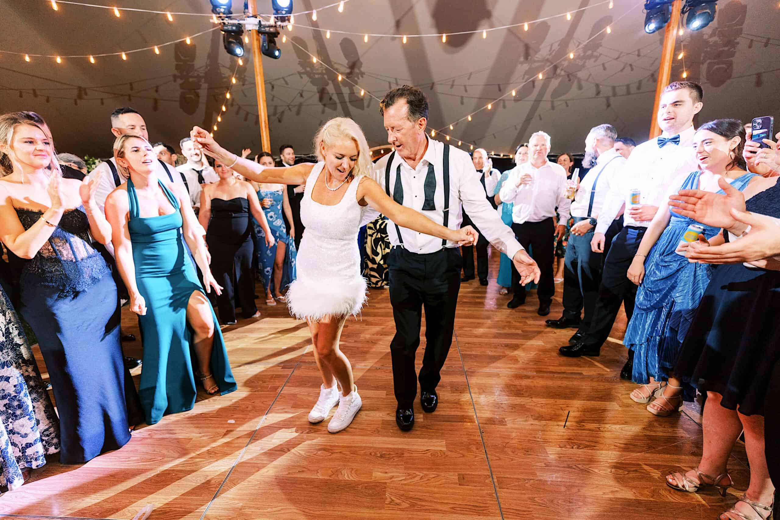 At a romantic backyard wedding in NJ, a woman in a white dress and sneakers dances with an older man in suspenders on a wooden floor, surrounded by clapping guests under string lights.